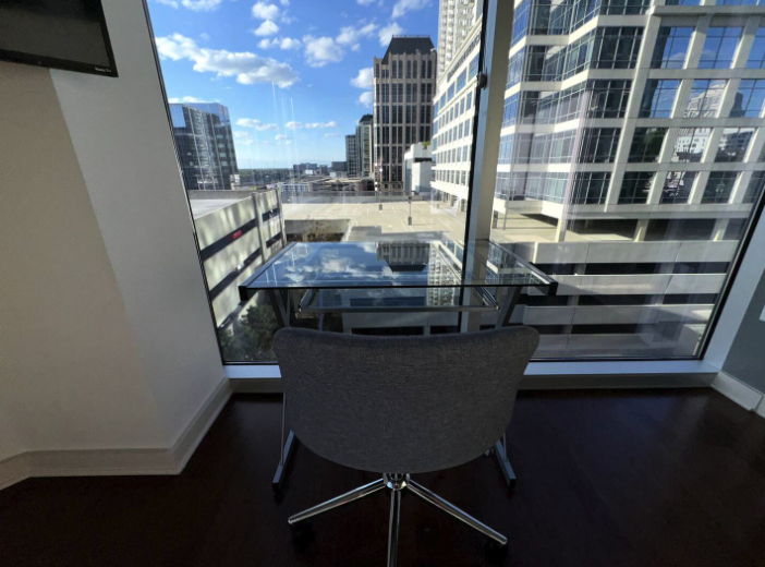 A small office space with a glass table and a gray office chair facing a floor-to-ceiling window. The window offers a view of tall modern buildings and a cityscape under a partly cloudy sky.