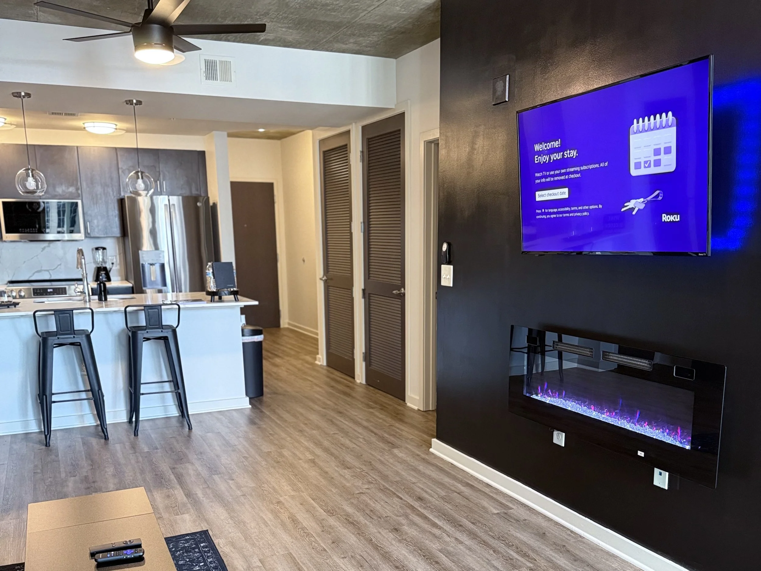Modern open-concept living space with kitchen, including a stainless steel refrigerator, microwave, and kitchen island with two barstools. To the right, a wall-mounted TV displays a welcome screen, with an electric fireplace beneath it. The area feat