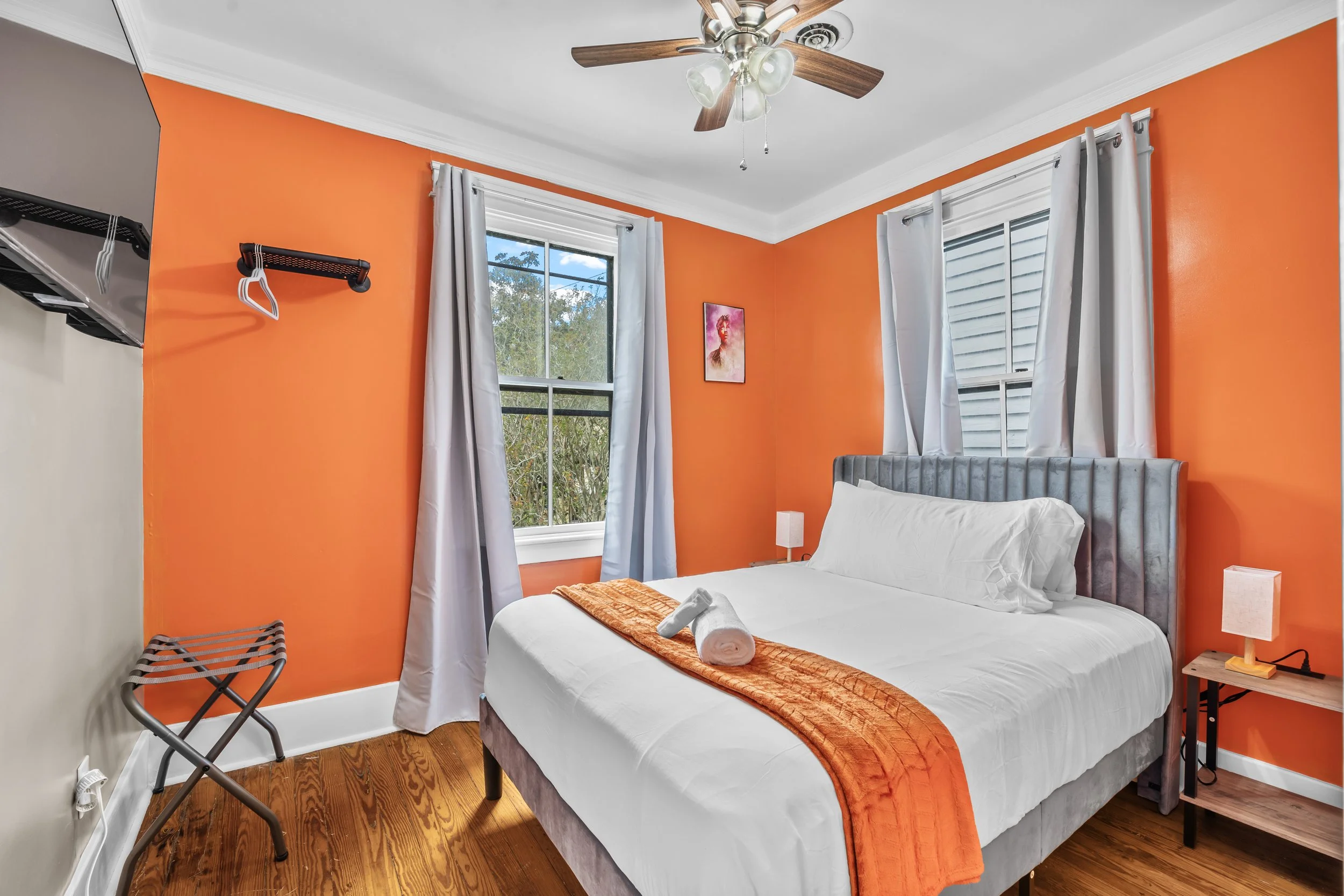 A bedroom with orange accent walls, a gray upholstered bed with white sheets, an orange runner, and a rolled towel on the bed. There are two windows with white curtains, a small wooden nightstand with a lamp, a ceiling fan with wooden blades, and a w