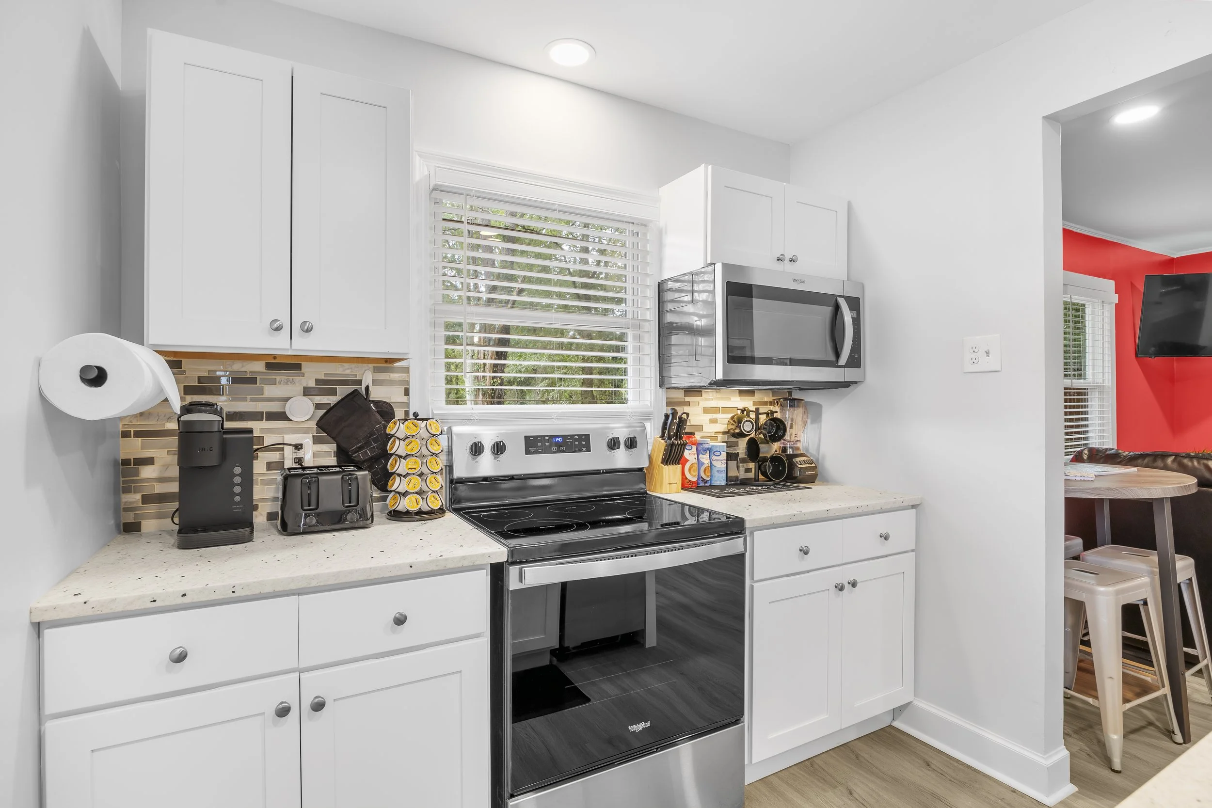 Modern kitchen with white cabinets, black stove, microwave, small appliances, and a window with blinds, adjacent to a dining area with a red wall and a mounted TV.