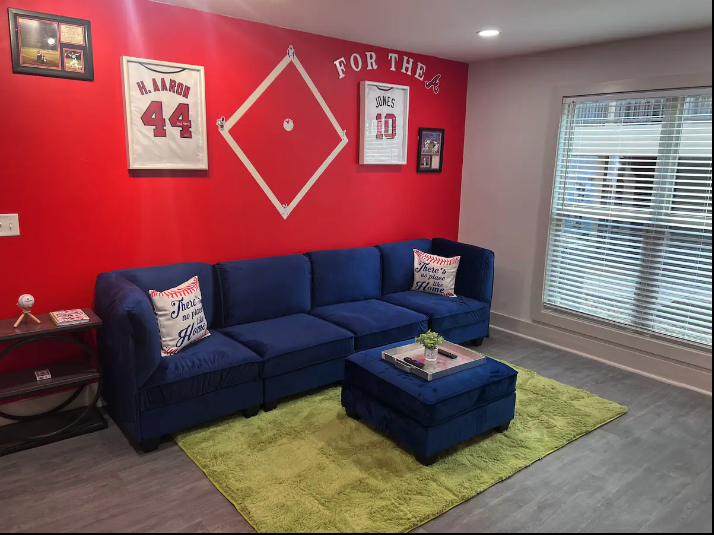 Living room with a red accent wall decorated with sports memorabilia, including framed jerseys of H. Aaron and Jones, and a baseball diamond-shaped frame. A blue sofa with decorative pillows sits on a green rug, with a matching blue ottoman. There is