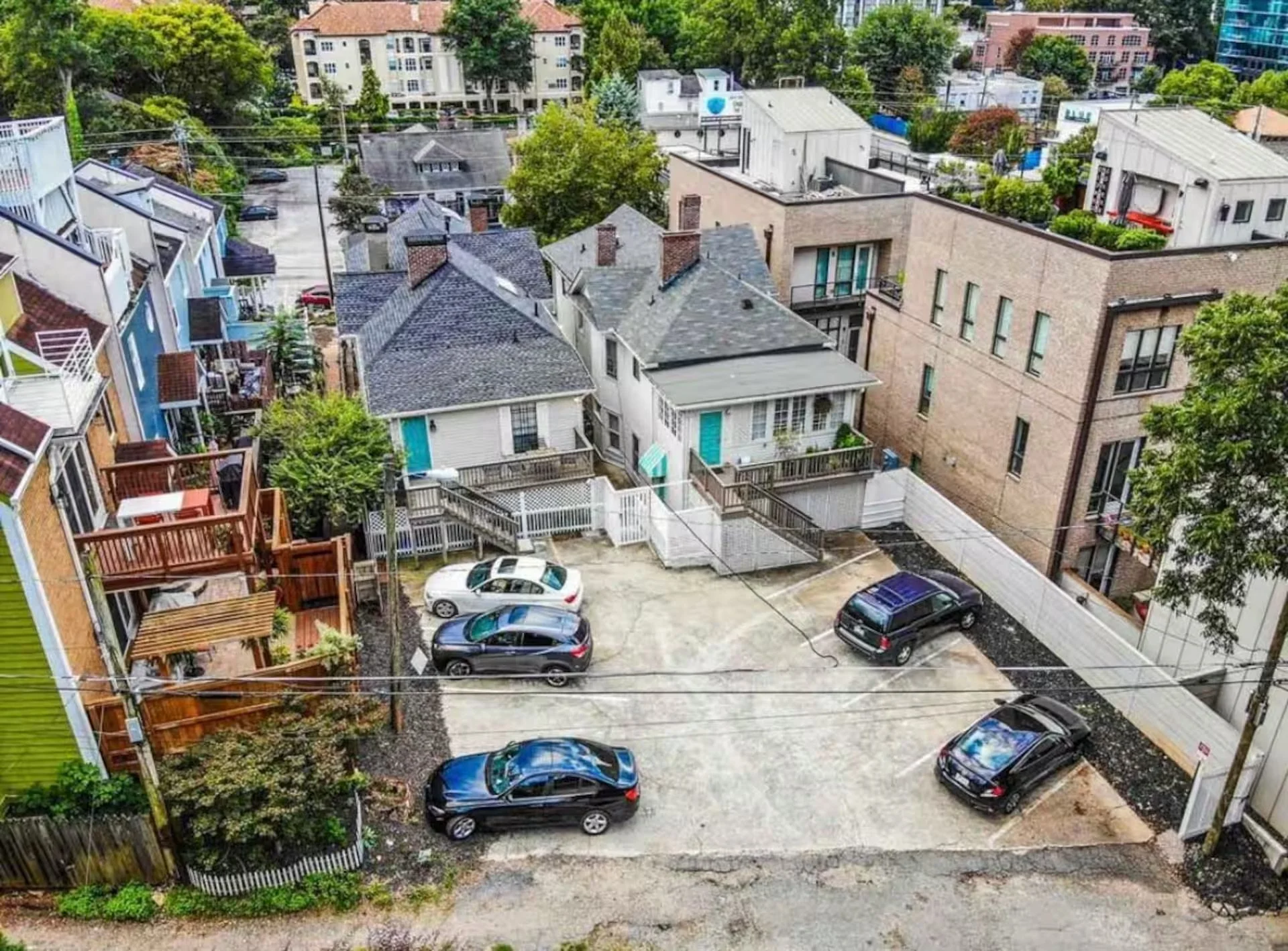A view from a balcony showing a parking lot with several cars, surrounding residential buildings, and green trees.
