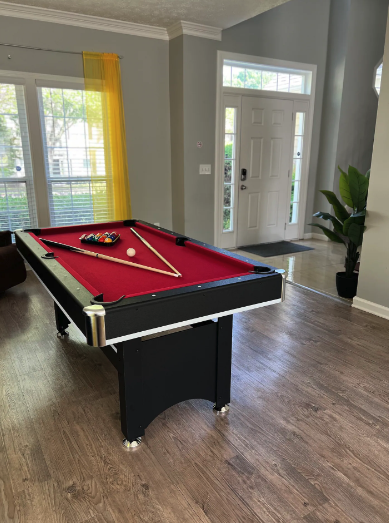 A black and red pool table with a cue stick, chalk, and white ball inside a living room near a white front door and a window with yellow curtains.