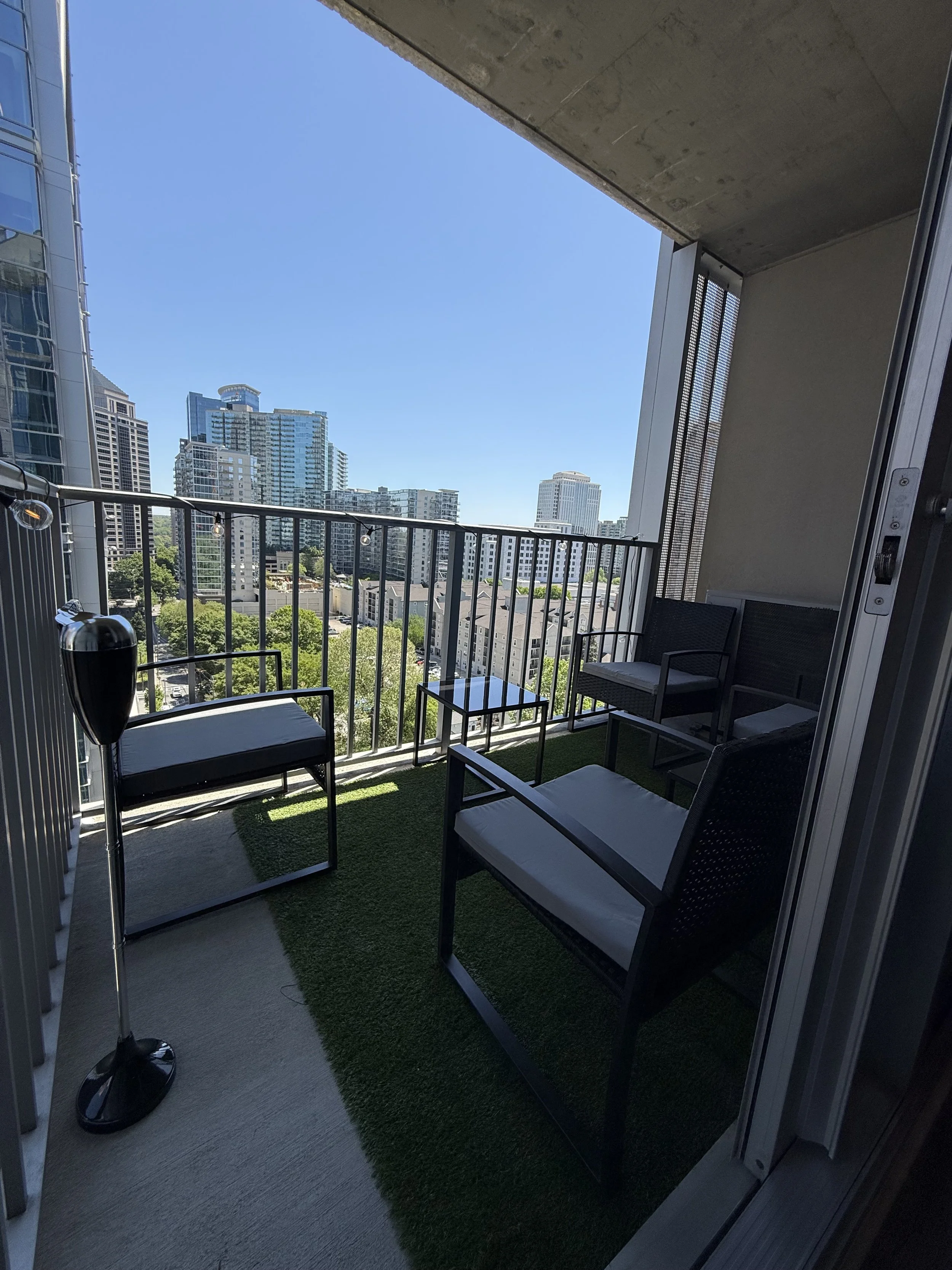 Balcony with black outdoor furniture, small table, artificial grass, city skyline with tall buildings, blue sky.