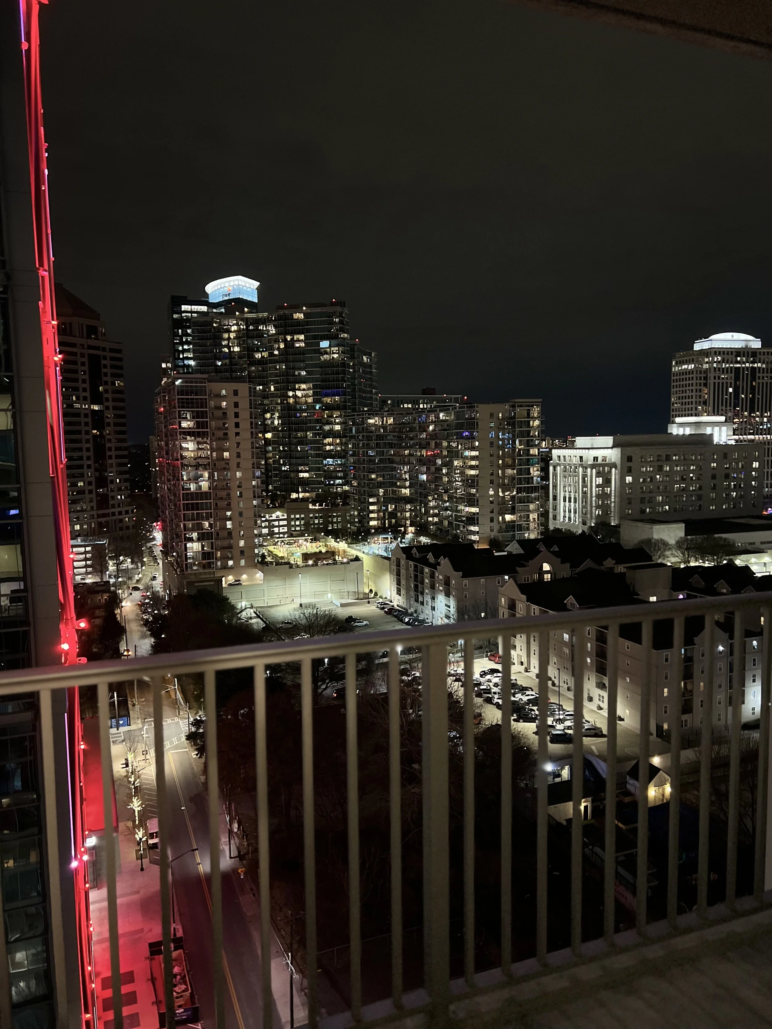 Nighttime cityscape view from a balcony showing tall buildings, lit windows, and a street below with parked cars and streetlights.