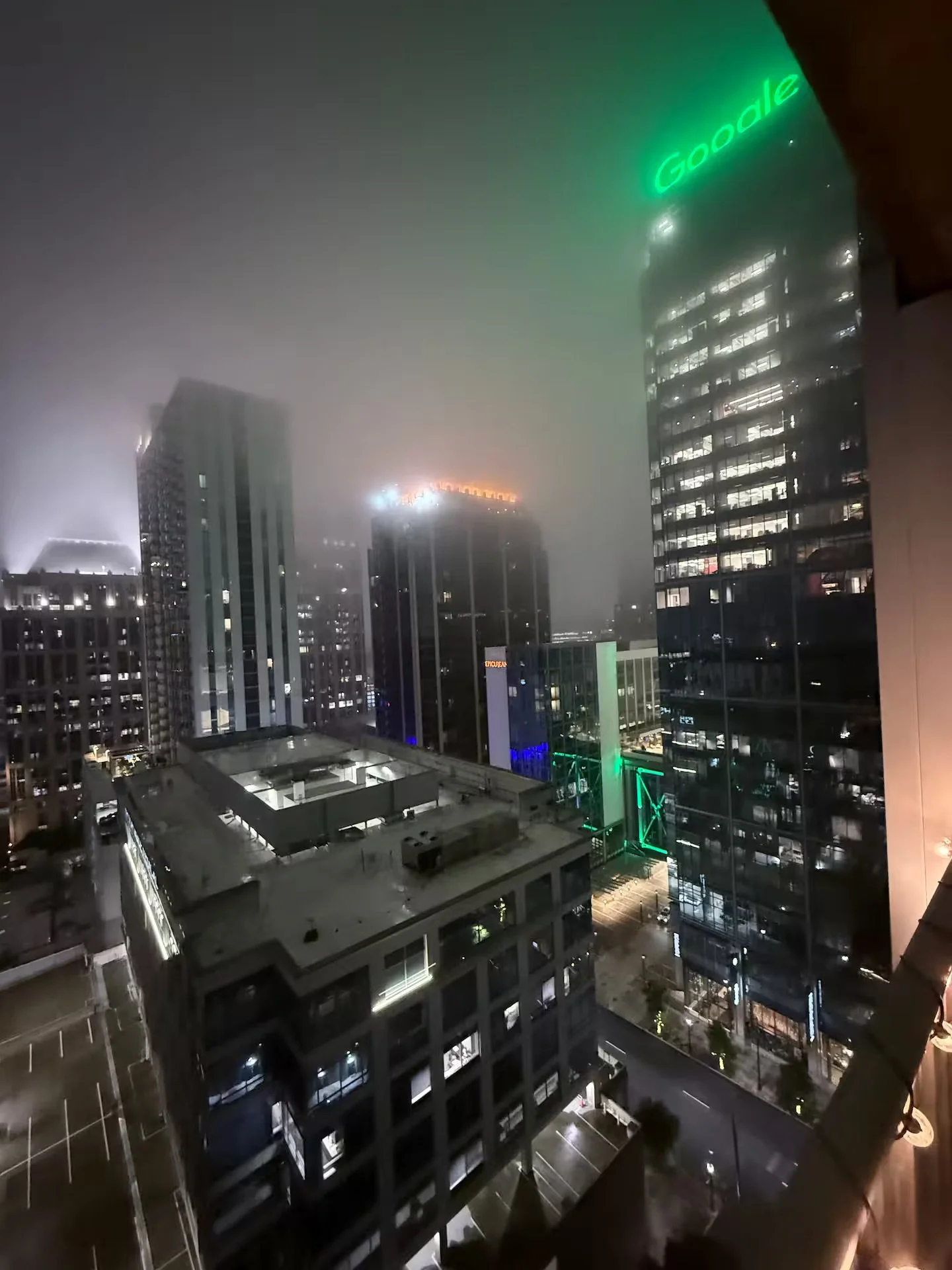 Nighttime cityscape with fog, illuminated skyscrapers, some with lights on, and signs including a prominently visible green neon 'Google' sign on a tall building.