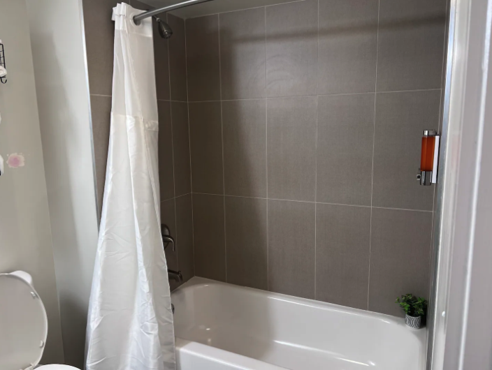 Bathroom with a white bathtub, beige tiled wall, white shower curtain, small potted plant on the edge of the tub, and a wall-mounted orange soap dispenser.