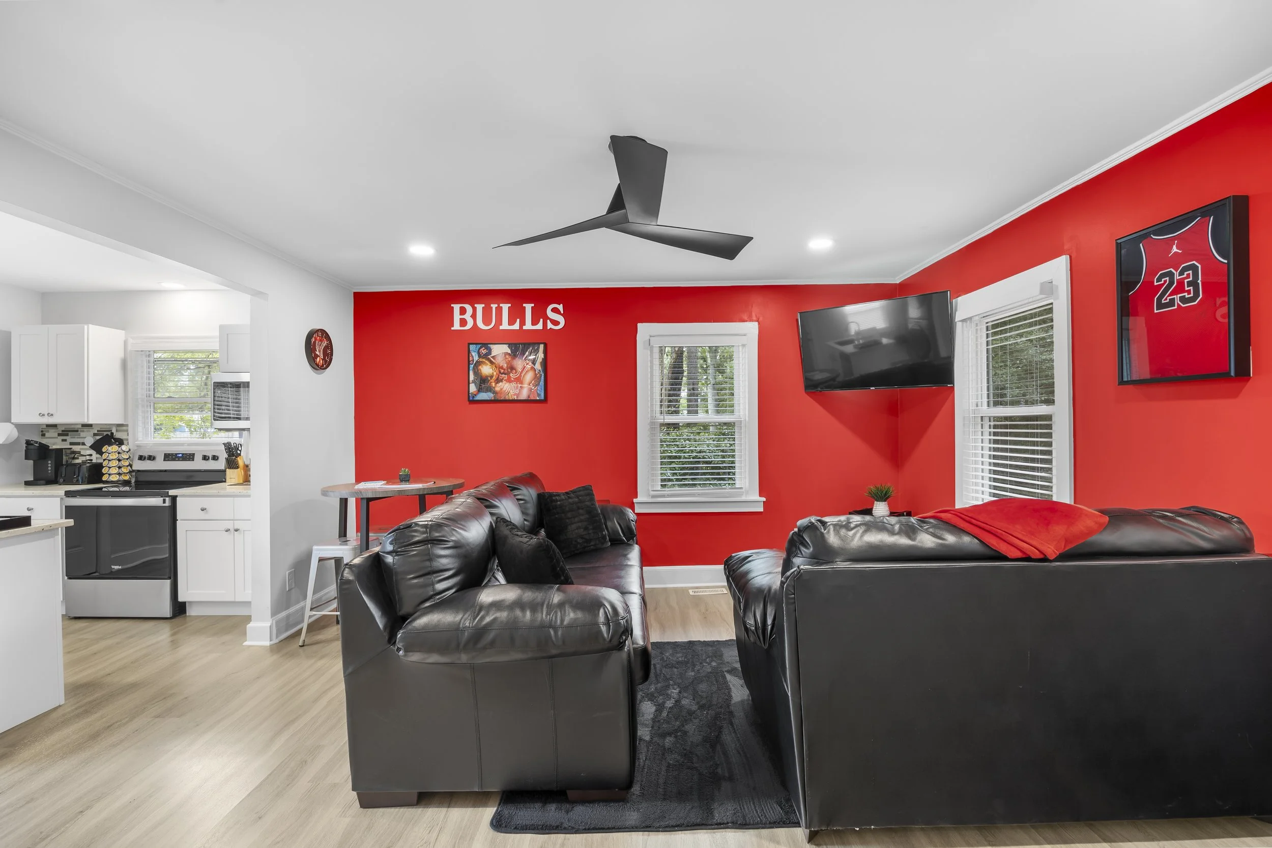 Living room with red accent wall, black leather sofas, wall art of basketball players, and a mounted flat-screen TV, with windows and a ceiling fan.