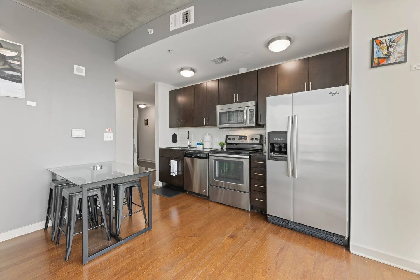 Modern kitchen with stainless steel appliances, dark wood cabinets, white walls, hardwood flooring, small table with stacked metal chairs, and abstract artwork.