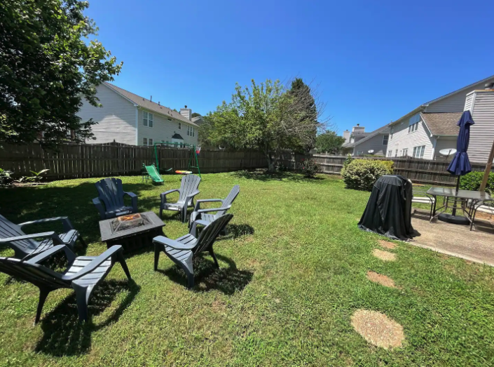 Backyard with outdoor furniture, a fire pit, a BBQ grill covered, and a concrete patio with an umbrella, surrounded by a wooden fence and neighboring houses, under a clear blue sky.