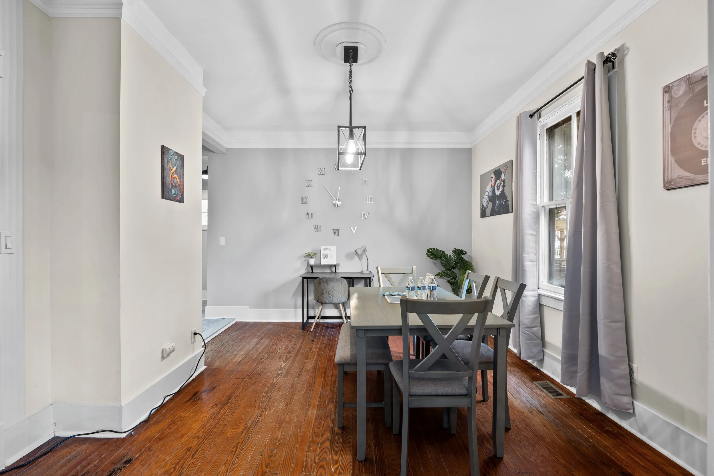 A dining room with gray walls, a wooden floor, a rectangular table with six chairs, a window with gray curtains, and wall art including a portrait and colorful artwork.