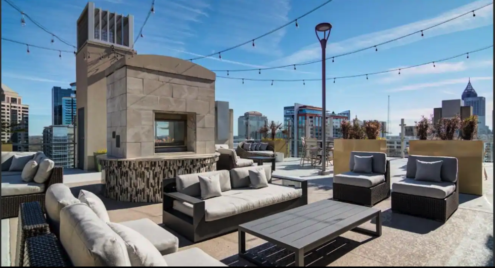 Rooftop lounge with sofas, armchairs, and tables, overlooking city skyline on a sunny day.