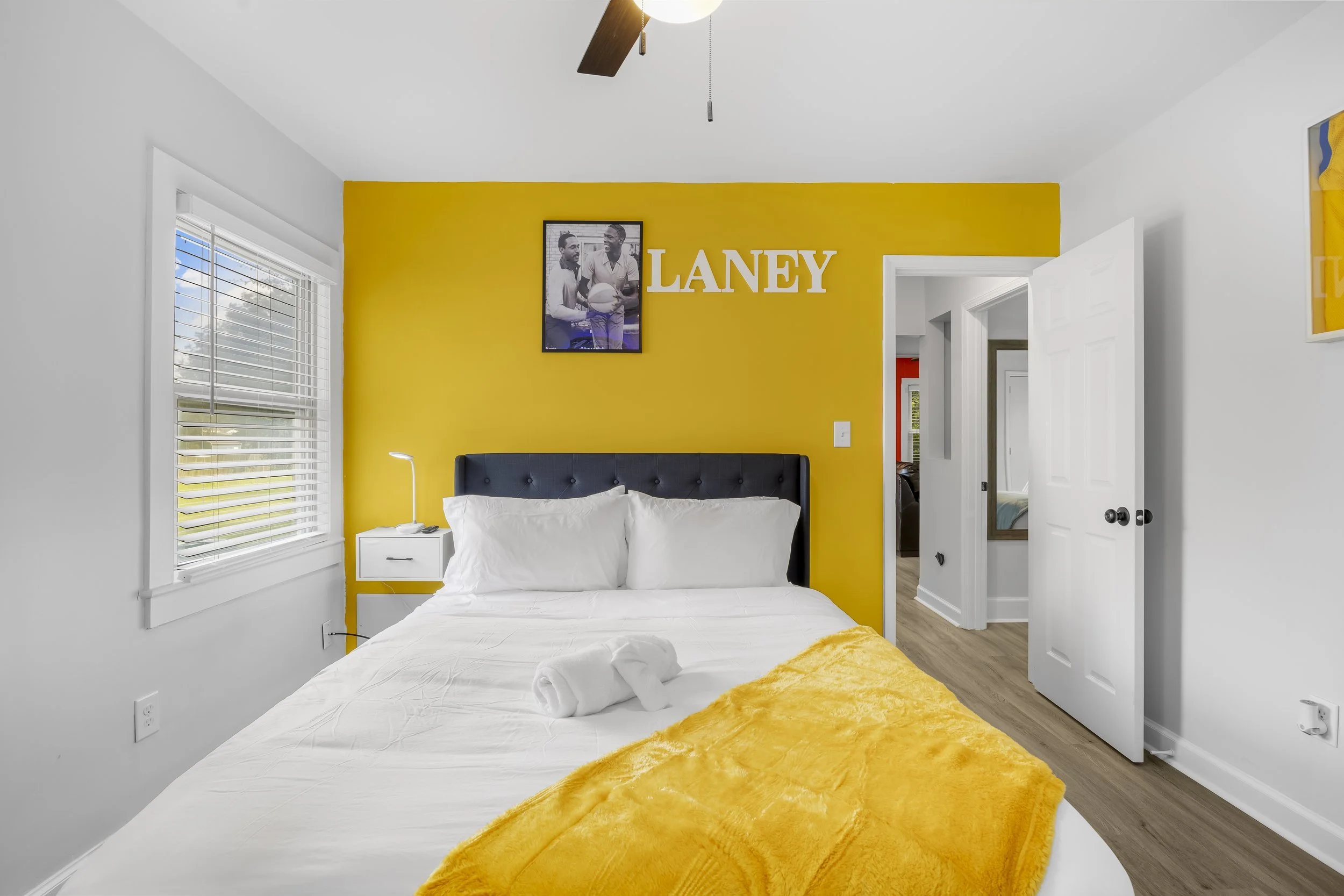 Bedroom with white walls and a yellow accent wall, black headboard, white linens, yellow blanket, window with blinds, nightstand with lamp, and a framed picture with the name 'LANEY' on the yellow wall.