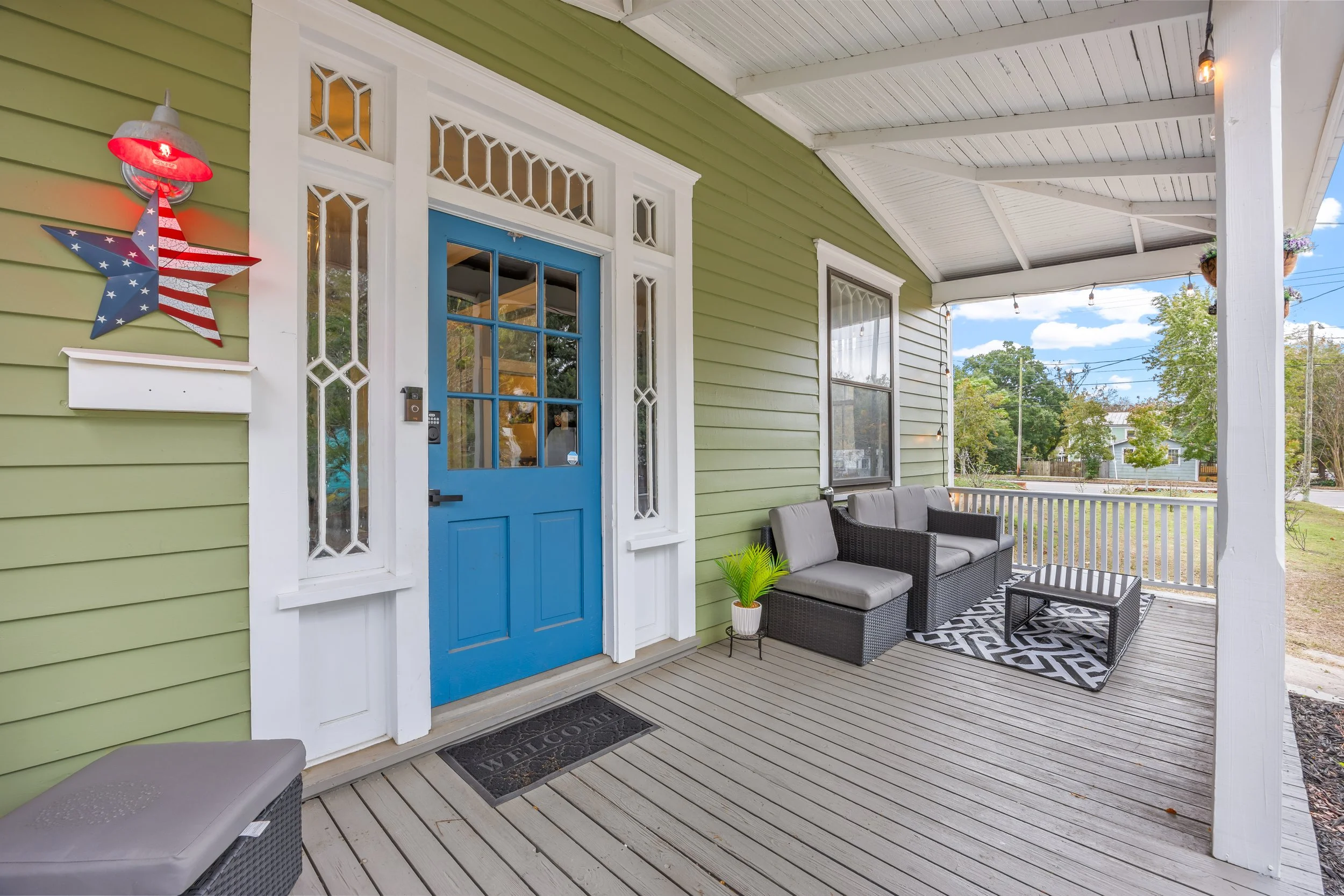Front porch of a house with a blue door, green siding, and white trim. There is outdoor furniture including a gray couch, black chair, and black coffee table on a wooden deck. Decor includes a patriotic star sign, a green potted plant, and a 'Welcome