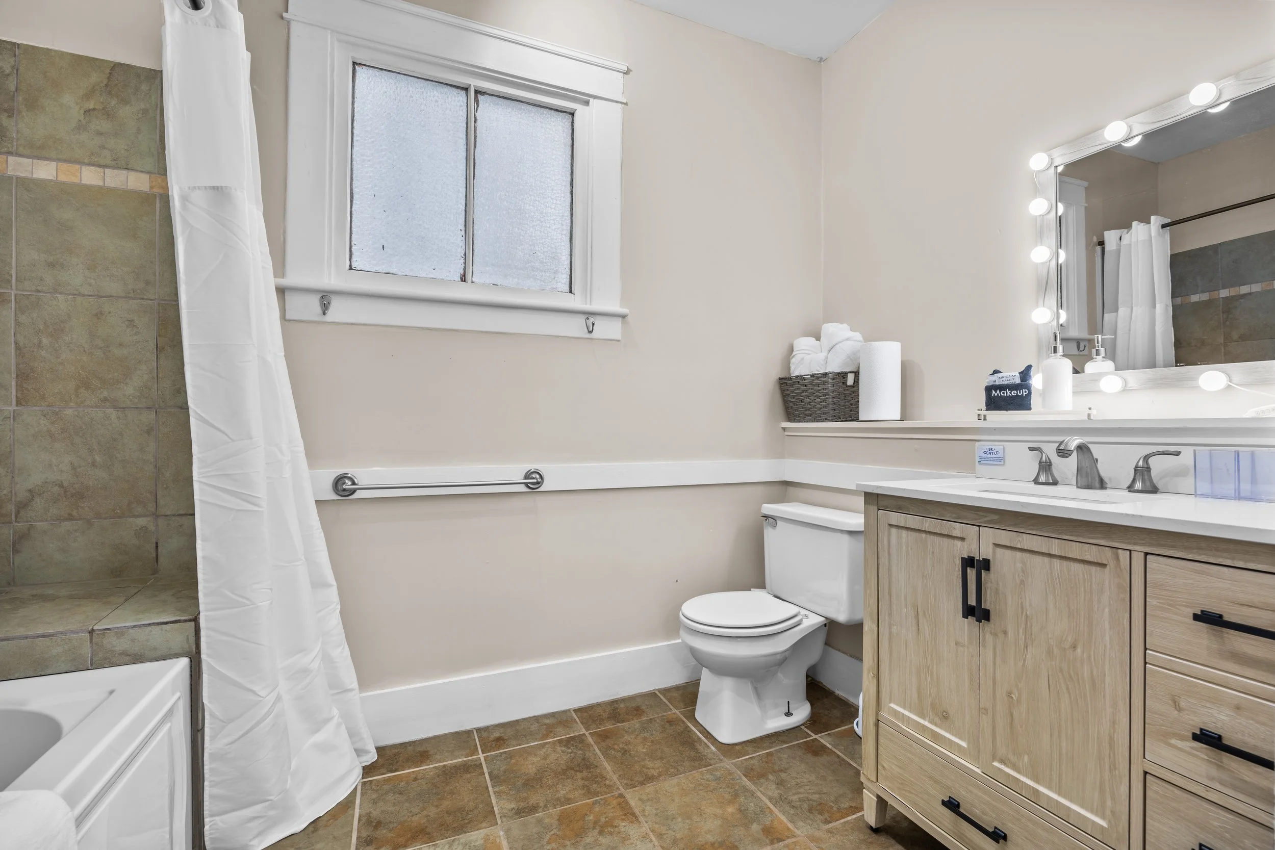 Bathroom with beige walls, a window, a toilet, a vanity with a mirror surrounded by lights, a basket of towels, and a shower with a curtain.