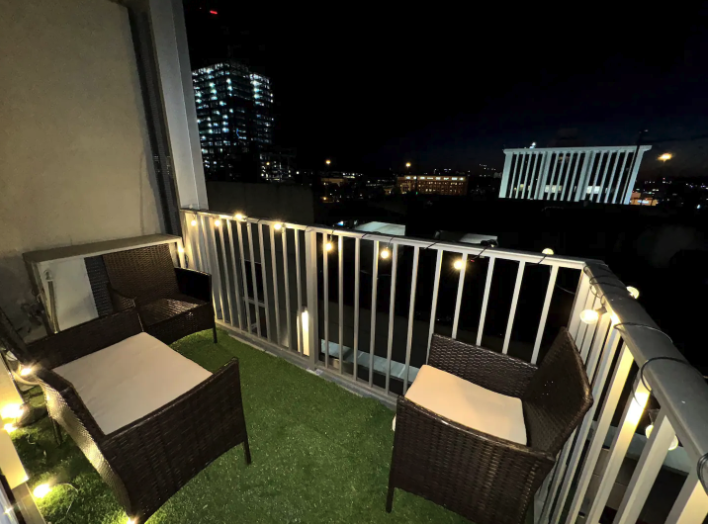 Nighttime view of a small outdoor balcony with four wicker chairs with cushions, string lights, artificial grass flooring, and city skyline with tall buildings in the background.