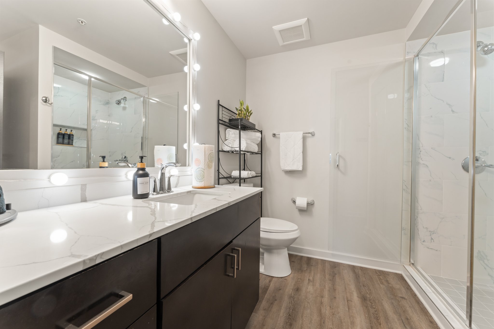Modern bathroom with a white marble countertop vanity, large mirror with bright lights, black cabinet, towel rack, and a glass-enclosed shower with marble tiles on the wall. There is a toilet and black shelving with folded towels and a plant.