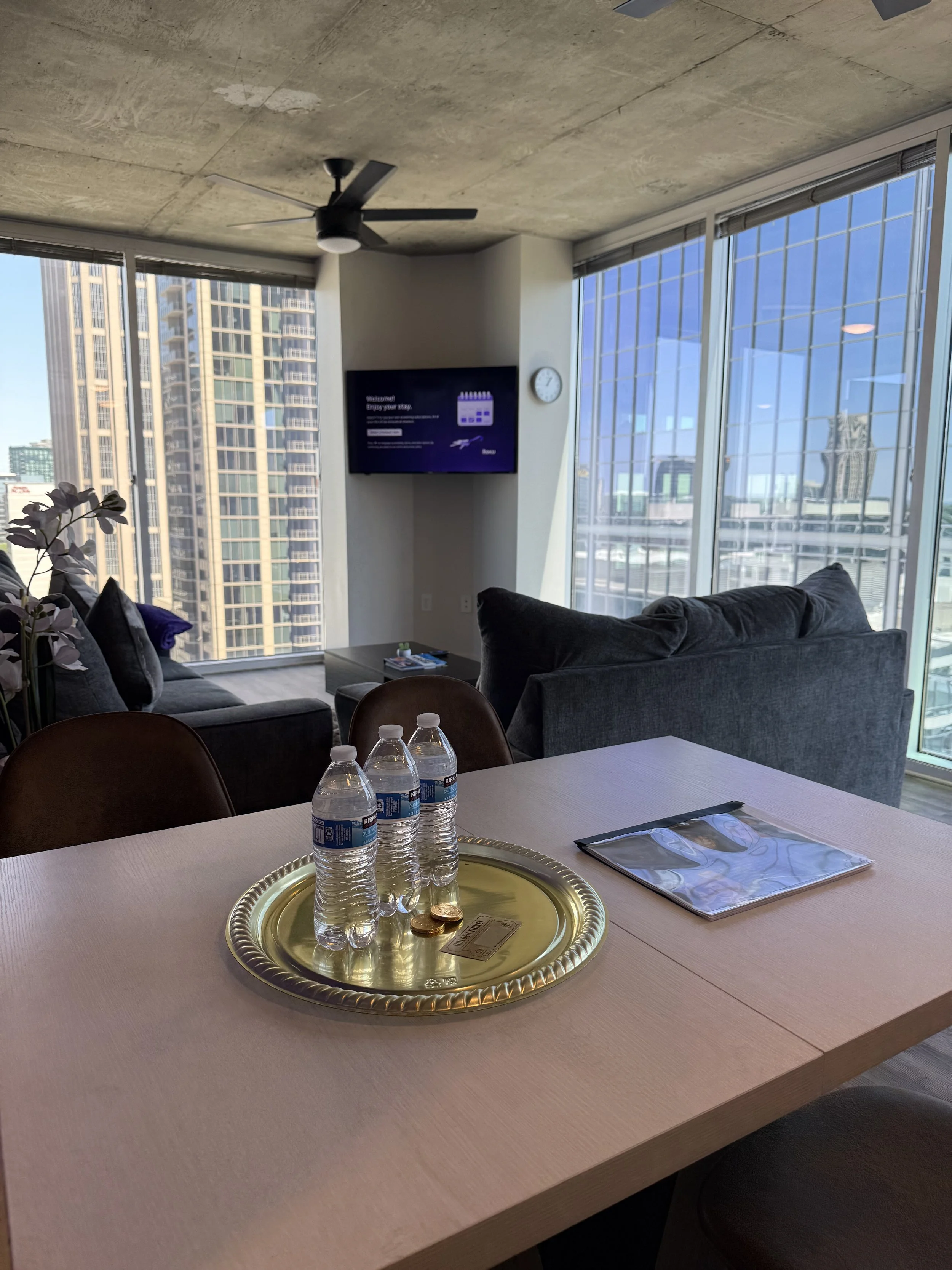 Modern living room with floor-to-ceiling windows showing city skyscrapers, a gray sofa, a coffee table, a TV on the wall, and a gold tray with bottled water and coins on a dining table.