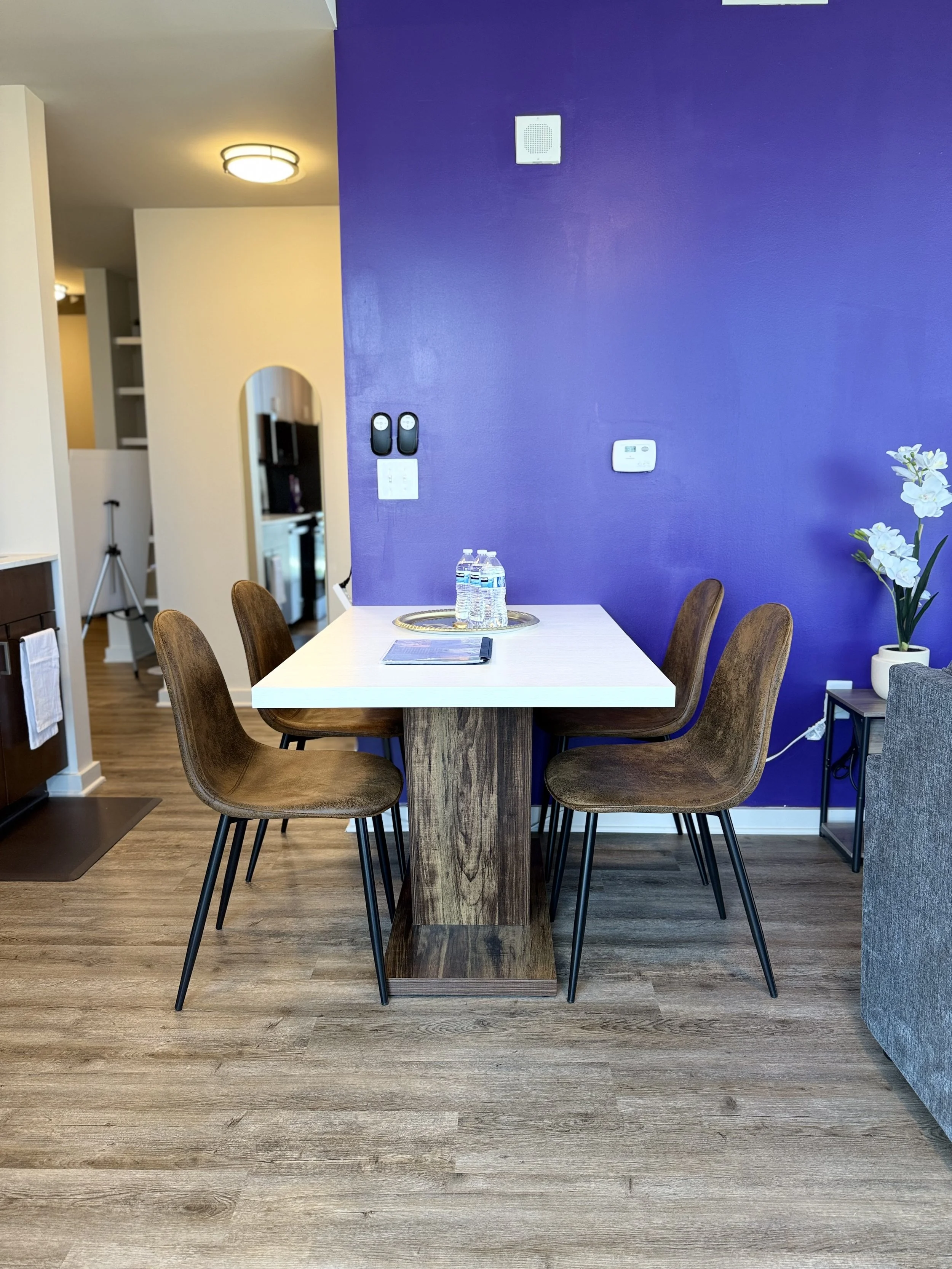 Dining table with four brown chairs, purple accent wall, bottles of water, remote, and a notepad, with a flower in a white pot on the side table.