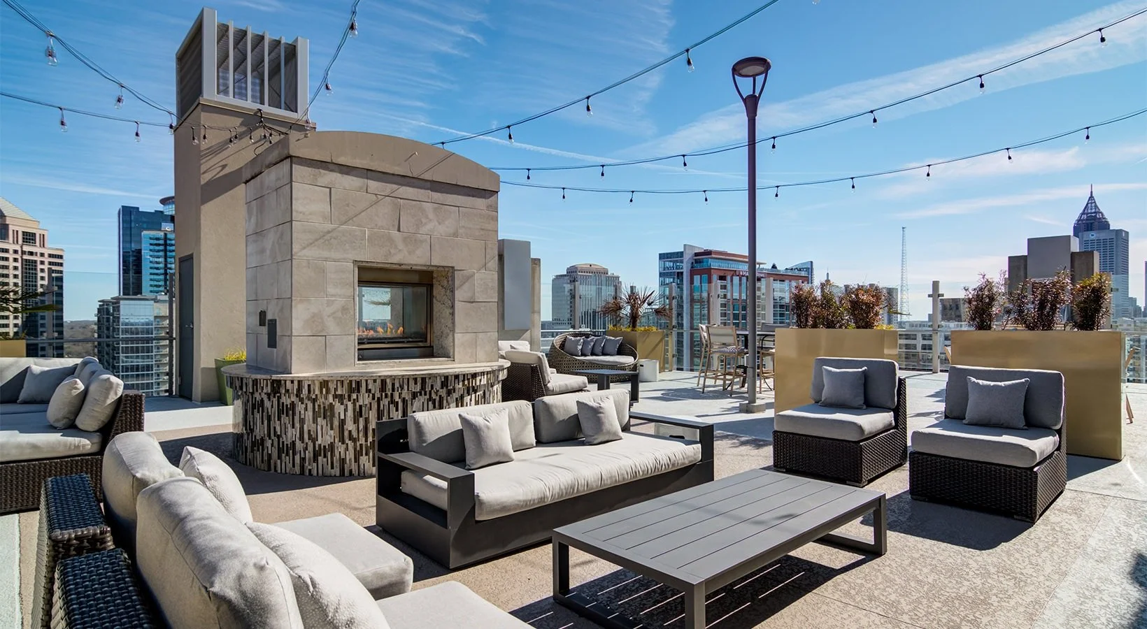 Rooftop lounge with outdoor seating, a fireplace, string lights, potted plants, and city skyline in the background.