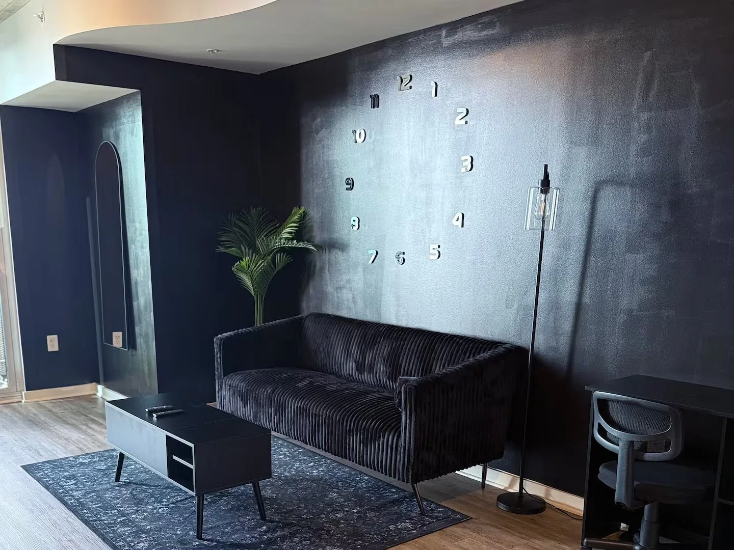 Modern living room with black textured wall, black velvet sofa, black coffee table, green plant, large wall clock with silver numbers, black standing floor lamp, and a black desk with a gray chair.