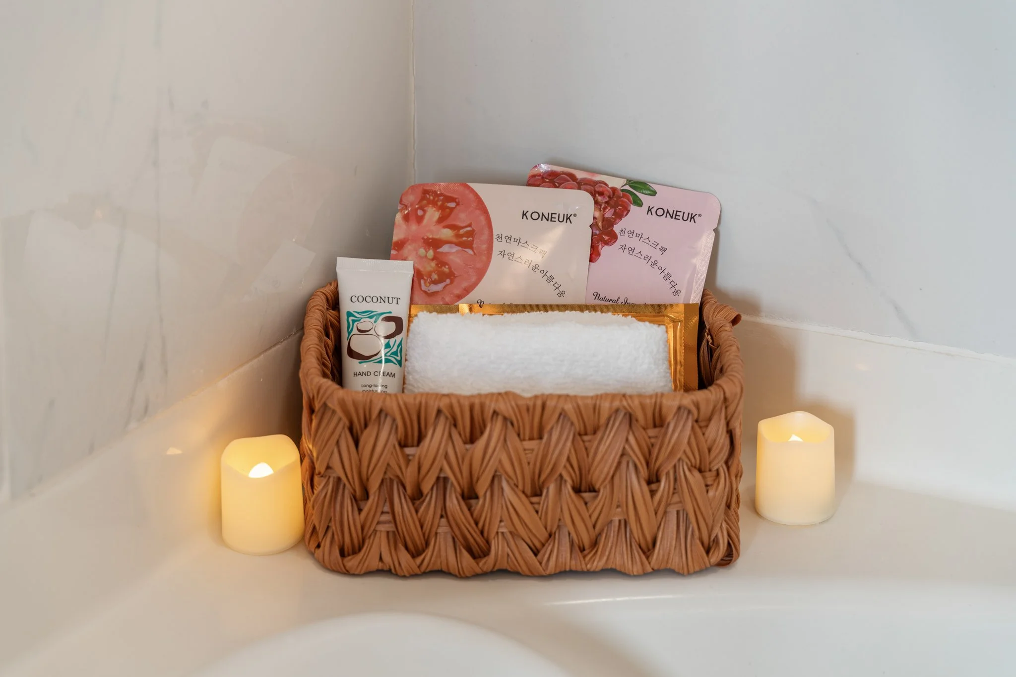 Wicker basket containing skincare products, including face masks, hand cream, towel, and two LED candles on a white surface in a bathroom.