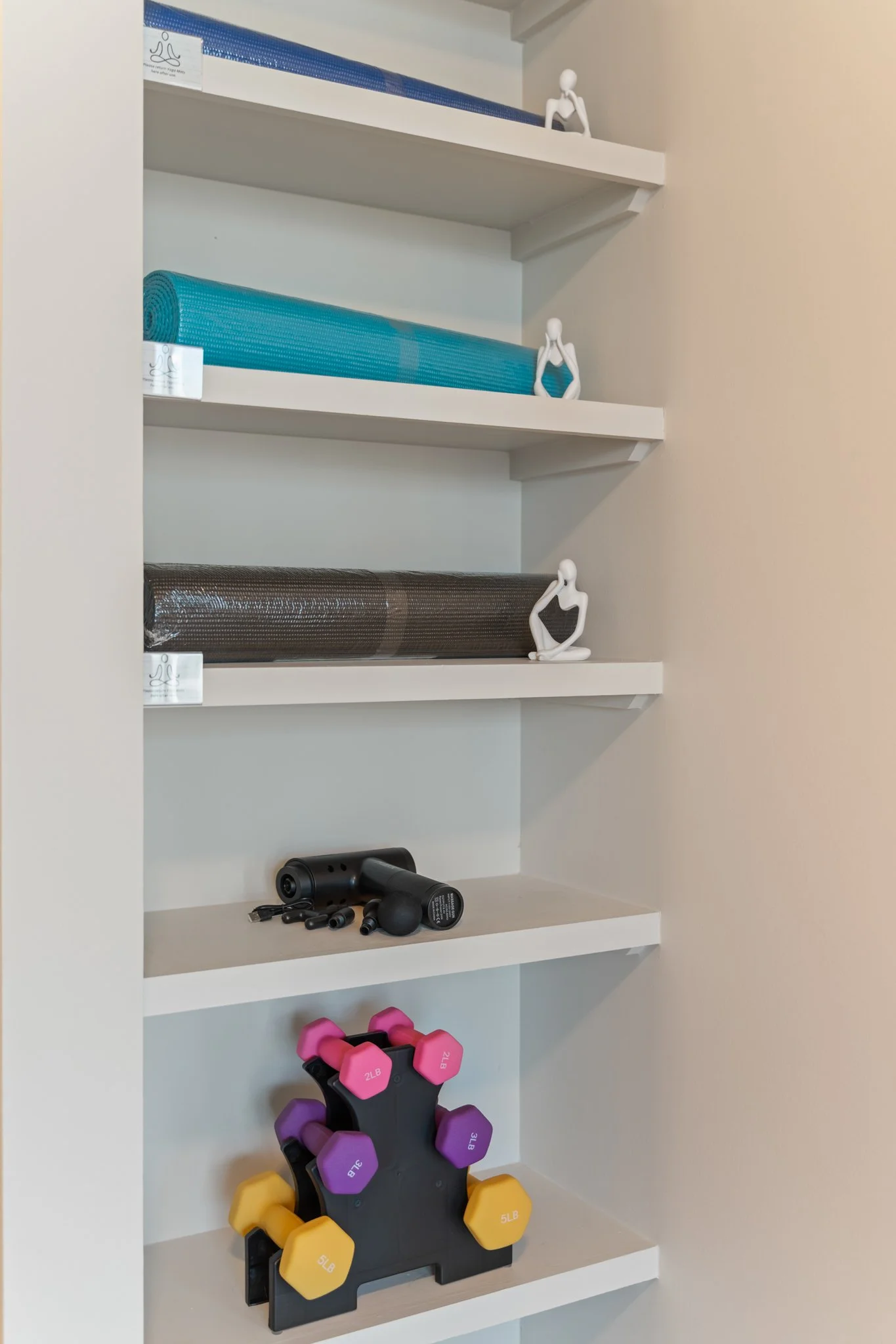 White shelves with exercise equipment, including yoga mats, a massage gun, and colorful dumbbells, along with small seated white figurines.