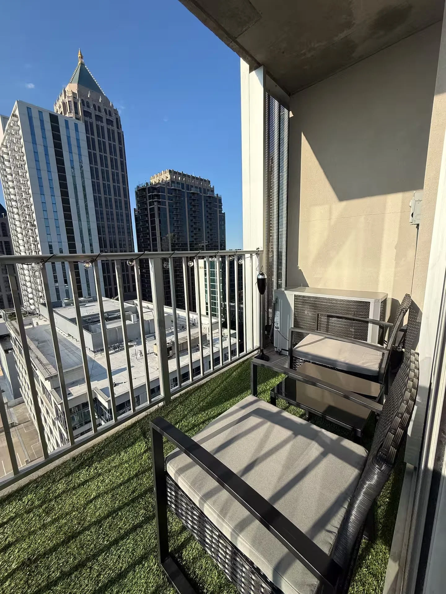 Small balcony with artificial grass flooring, two black metal chairs with cushions, string lights, and city skyscrapers in the background.