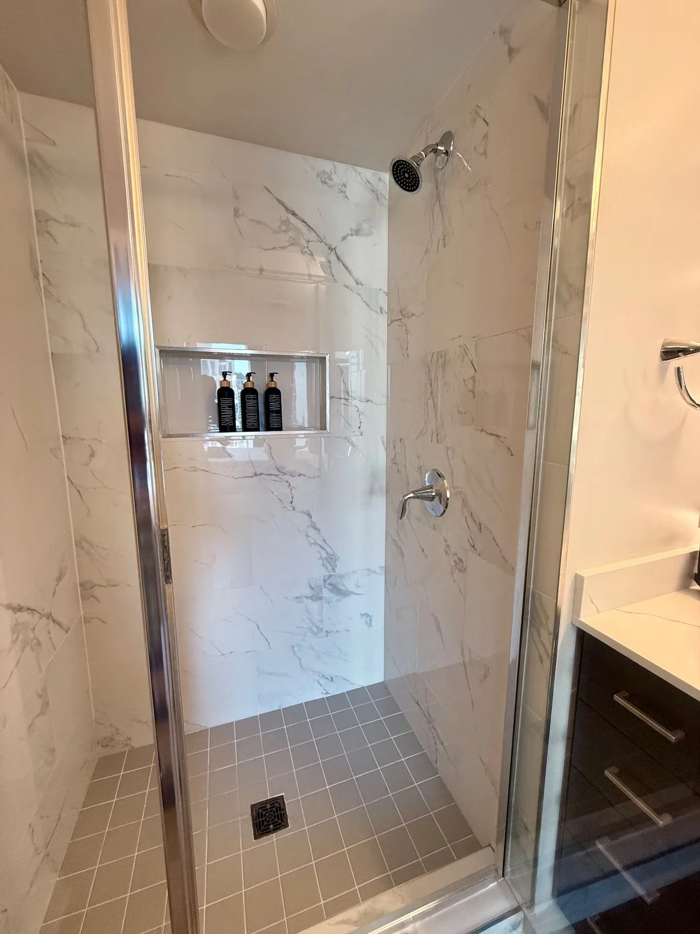 A walk-in shower with marble tile walls, a chrome showerhead, a built-in shelf with black bottles, and grey tiled flooring with a drain.