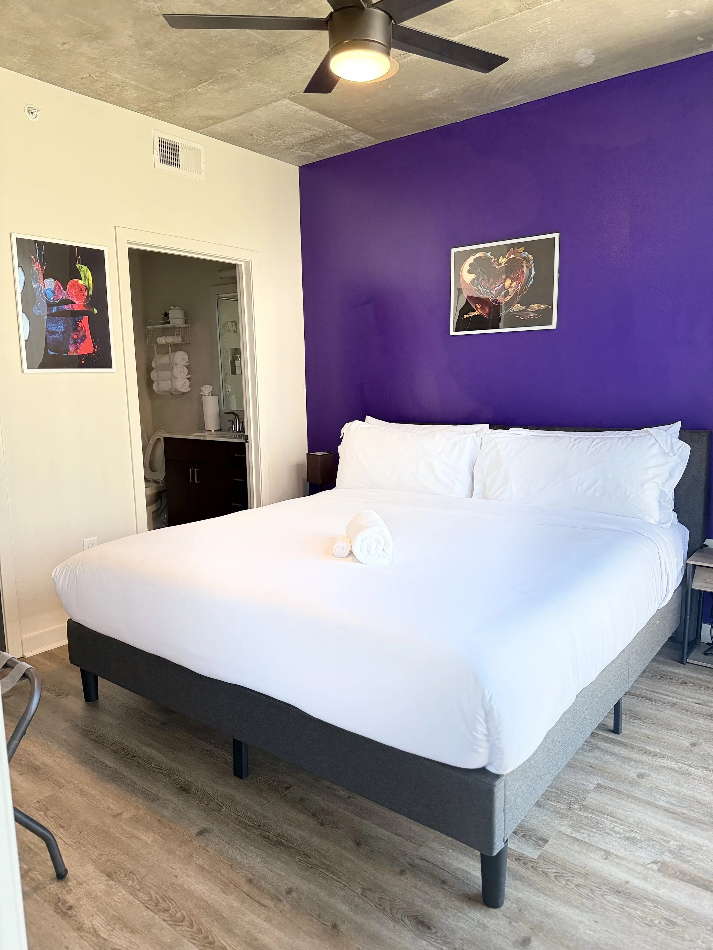A modern hotel room with a white bed against a purple accent wall, decorated with artwork, and a ceiling fan. There are two rolled towels on the bed, and a small bedside table on one side. A doorway leads to a bathroom visible in the background.