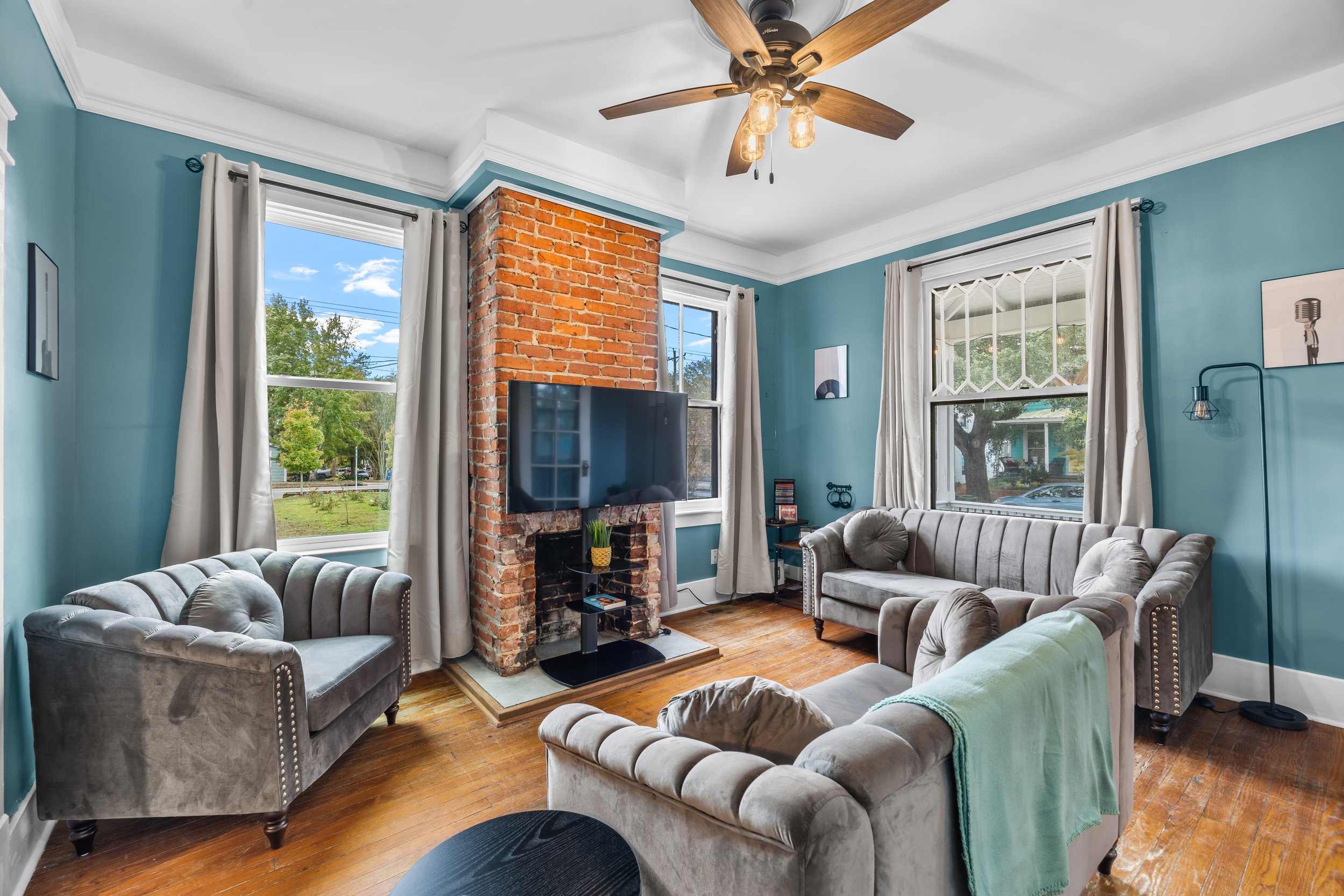 Living room with teal walls, white curtains, a brick fireplace with a mounted flat-screen TV, and beige velvet sofas arranged around a central area.