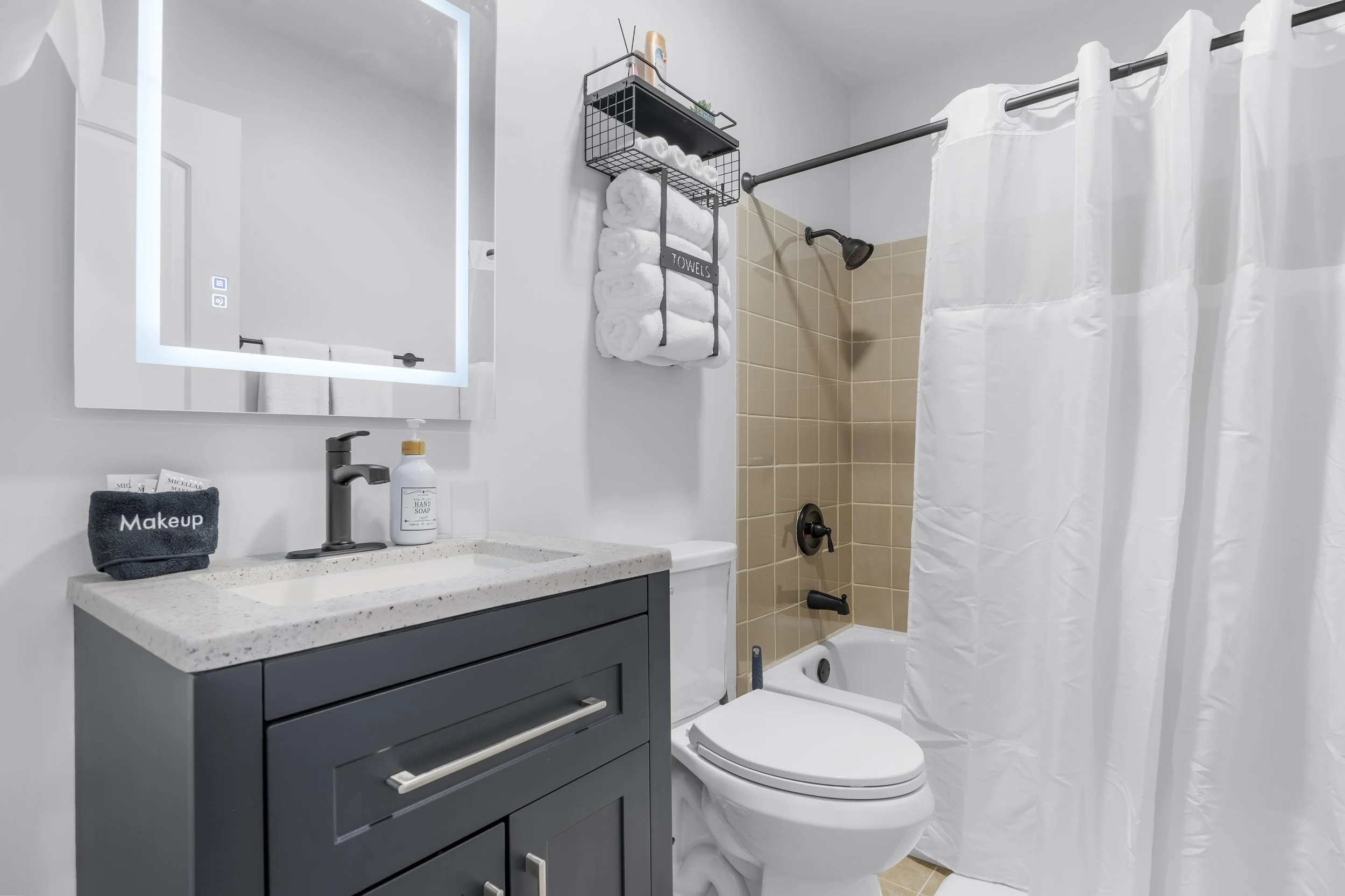 Modern bathroom with a navy vanity, white countertop, mirror with LED lighting, white towels in a wall-mounted rack, shower area with beige tiles, white shower curtain, and a black faucet.