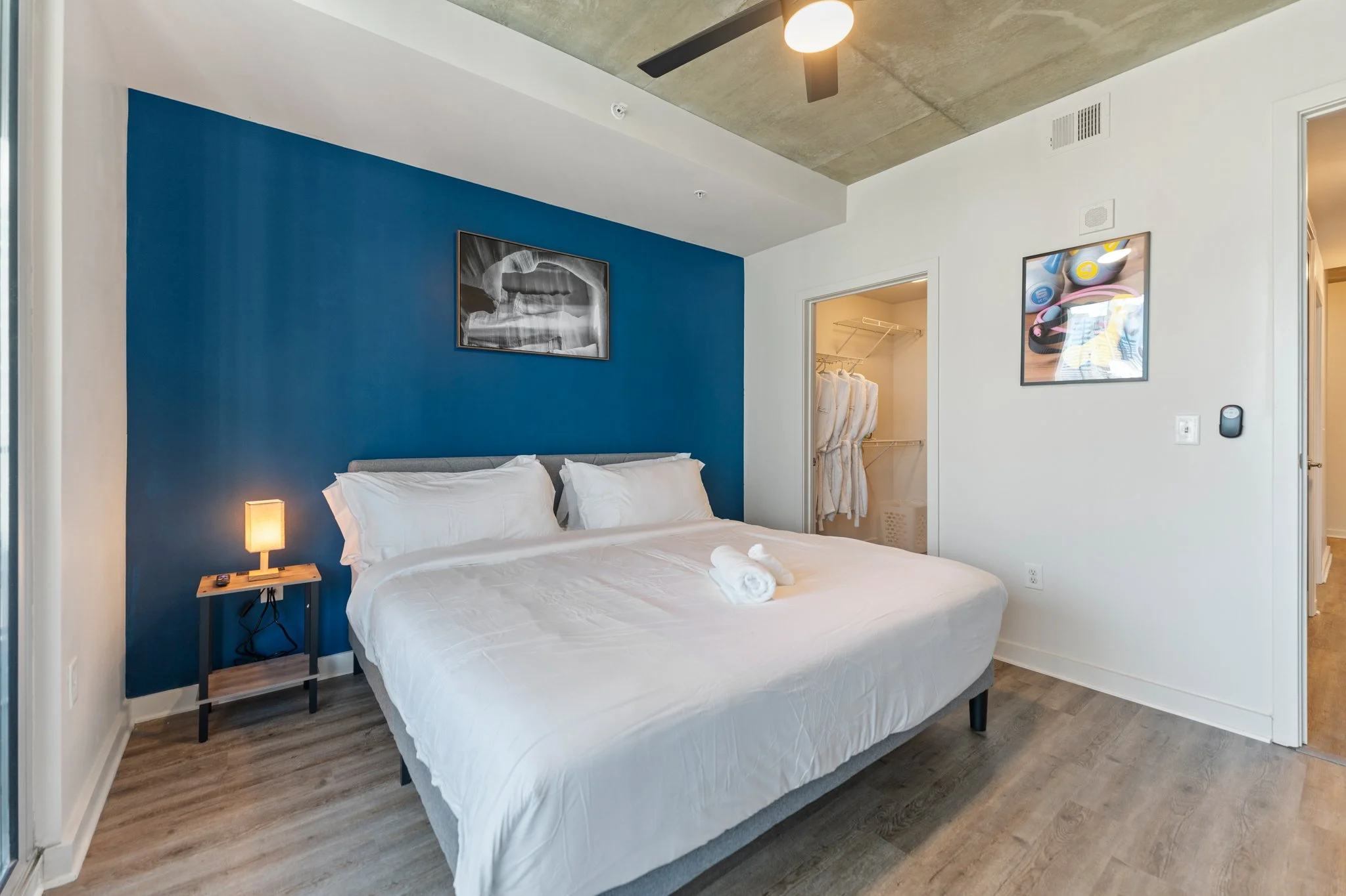 Modern bedroom with a white bed and blue accent wall, next to an open closet with hanging robes, a small nightstand with a lamp, and framed artwork on the walls.