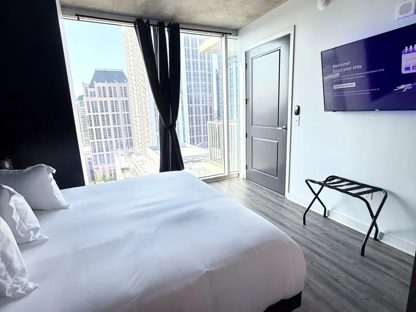A modern hotel room with a large bed, black curtains, a balcony door, a flat-screen TV mounted on the white wall, and a luggage rack, with city skyscrapers visible outside.