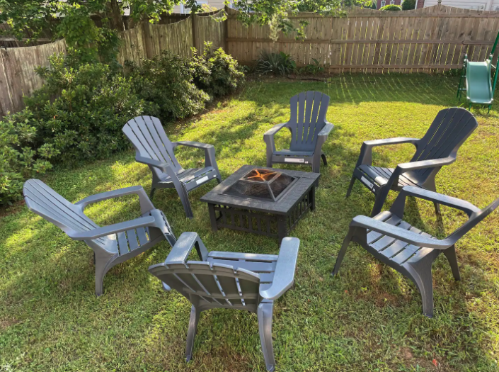 Six gray outdoor chairs arranged in a circle around a fire pit in a backyard lawn, with a wooden fence and trees in the background.