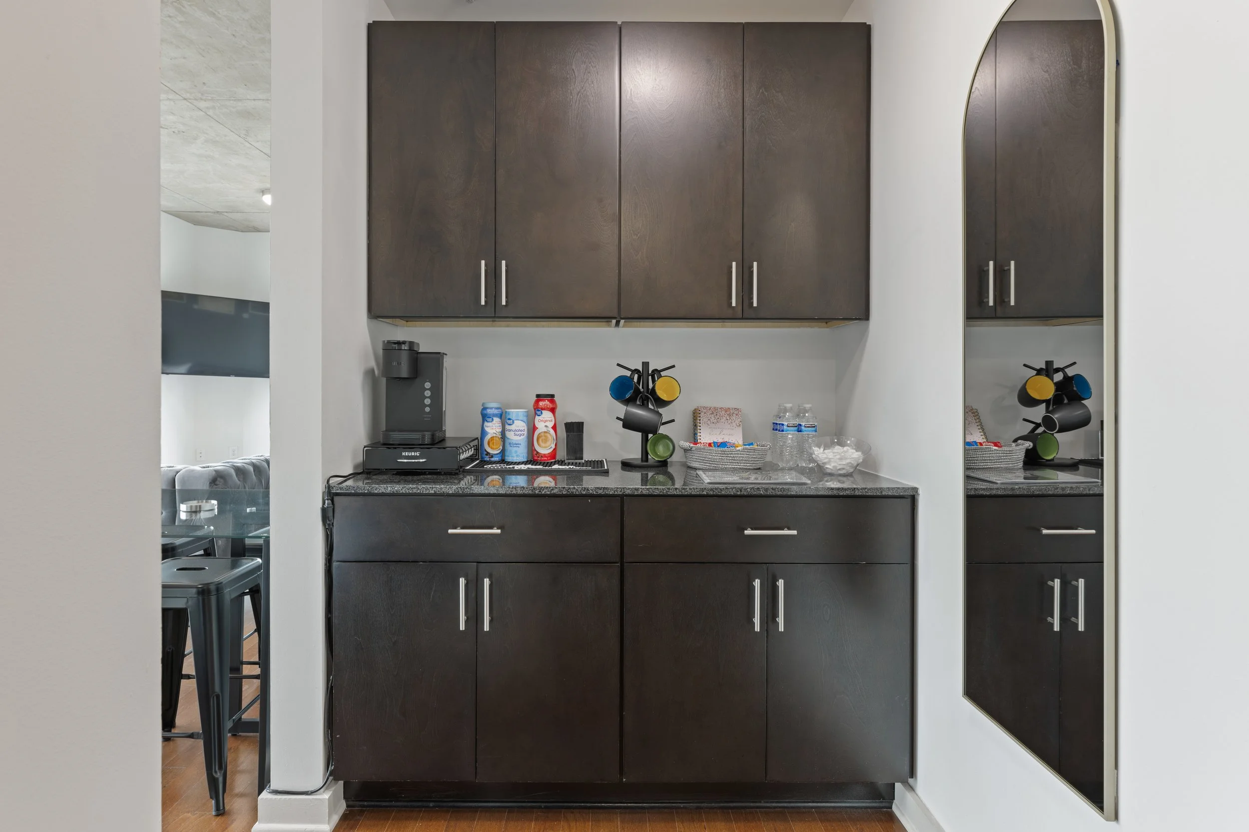 Kitchenette with dark wood cabinets, a coffee maker, drinks, cups in a mug holder, bottled water, and bowls, reflected in a tall mirror.