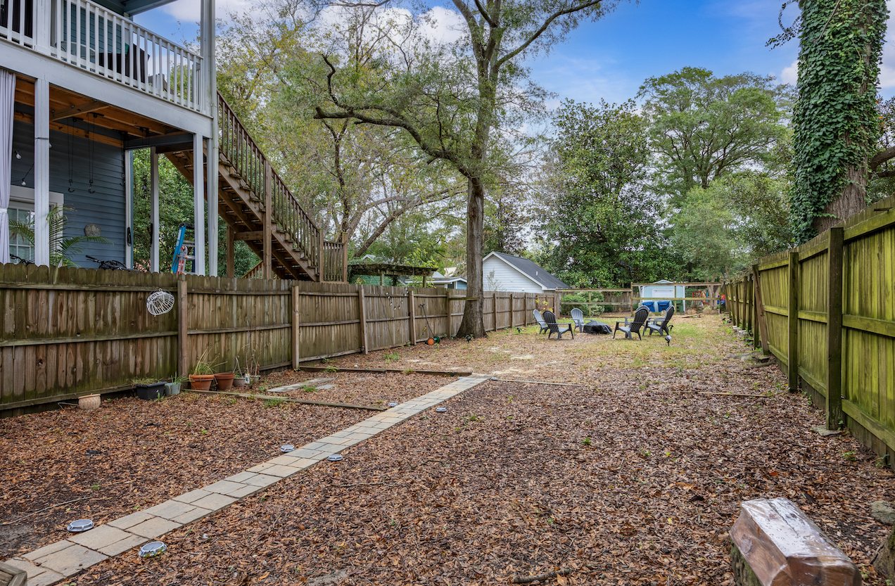 Backyard with leaf-covered ground, surrounded by wooden fences, featuring outdoor chairs, trees, and a staircase leading to a second-story porch of a blue house.