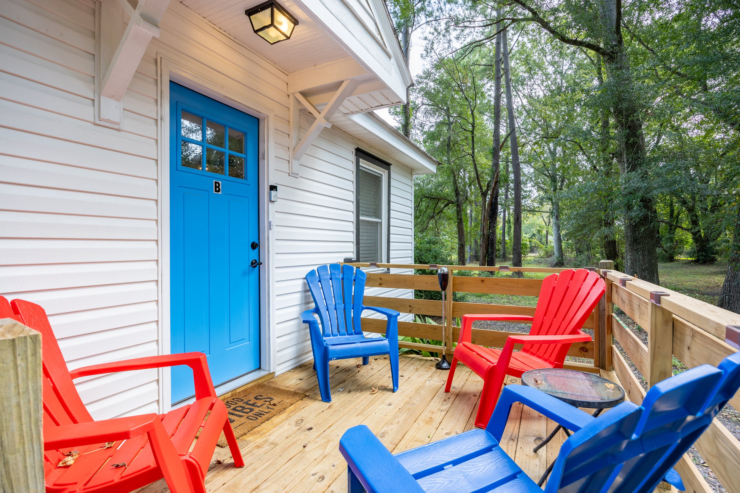 Front porch with white siding, blue door, and red and blue Adirondack chairs on a wooden deck, surrounded by trees.
