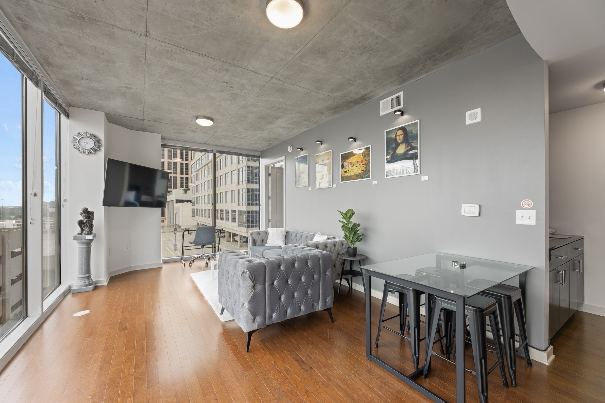 Modern high-rise apartment living room with gray walls, hardwood floors, a tufted gray sofa, a glass dining table with black stools, a wall-mounted TV, artwork on the wall, a potted plant, and large windows with city views.