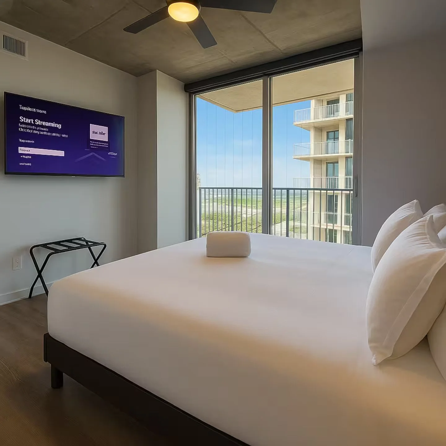 Modern bedroom with large bed, white bedding, pillows, towel, and a sliding glass door opening to a balcony with a view of neighboring building and landscape. Wall-mounted TV displays streaming app.