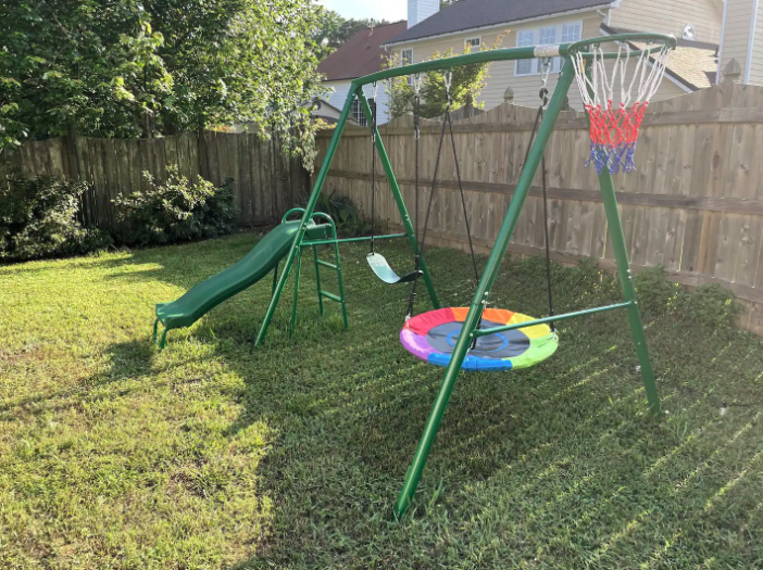 Green backyard playset with slide, swing, and basketball hoop, on grassy yard next to wooden fence.