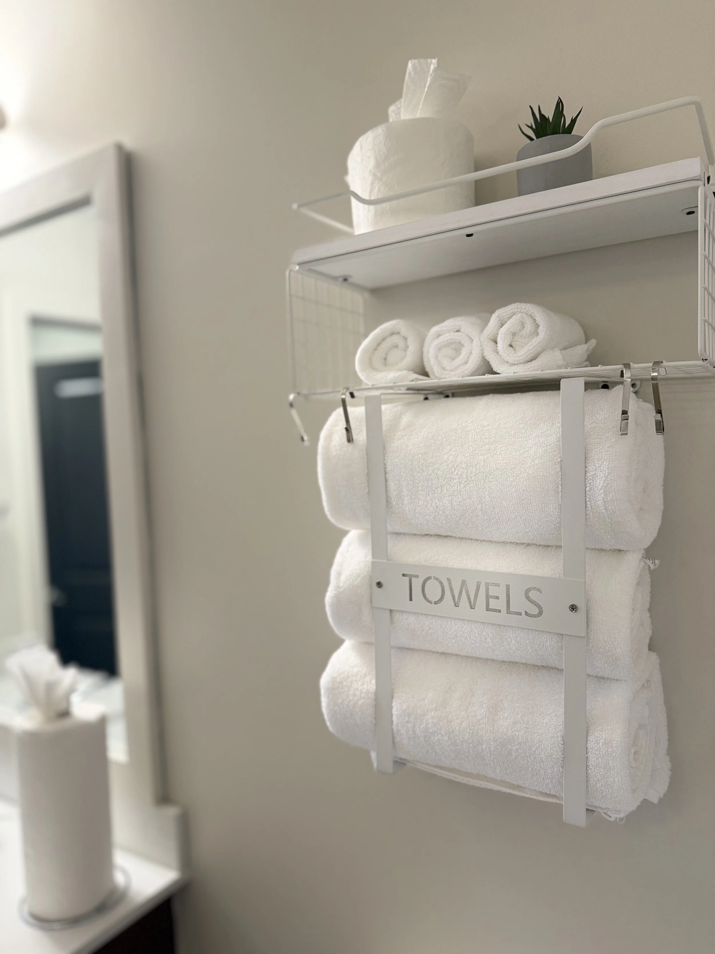 Wall-mounted white towel rack with rolled white towels and a sign that says 'Towels', top shelf with tissue paper and a small plant in a pot, background with a bathroom mirror and tissue dispenser.