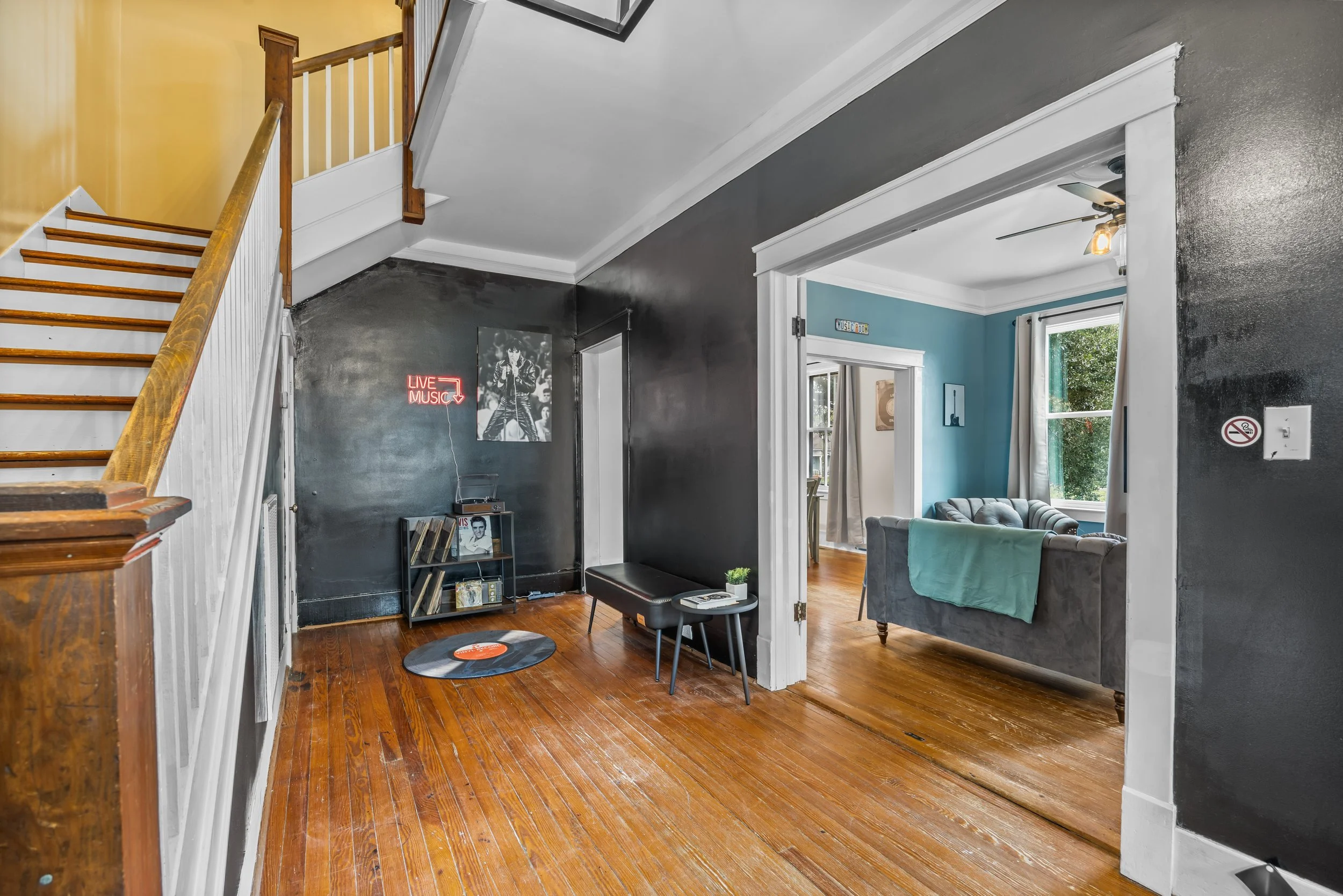 Living room with black and blue walls, wooden floor, and a staircase on the left. The black wall has a 'Live Music' sign, a picture of a singer, and a bookshelf with records. The adjacent room has a blue wall, a window with curtains, and a sofa with 