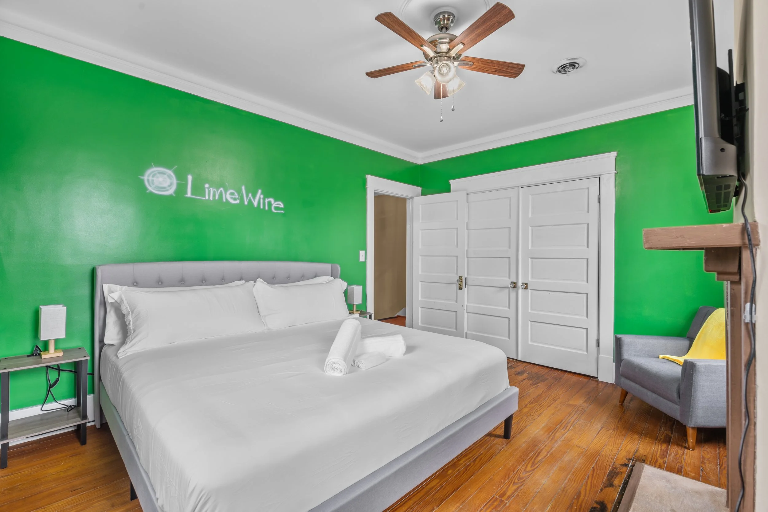 A bedroom with bright green walls, a gray upholstered bed with white linens and rolled towels, a small black nightstand with lamps, a ceiling fan with wooden blades, white double doors, a gray armchair with a yellow blanket, a mounted television, and