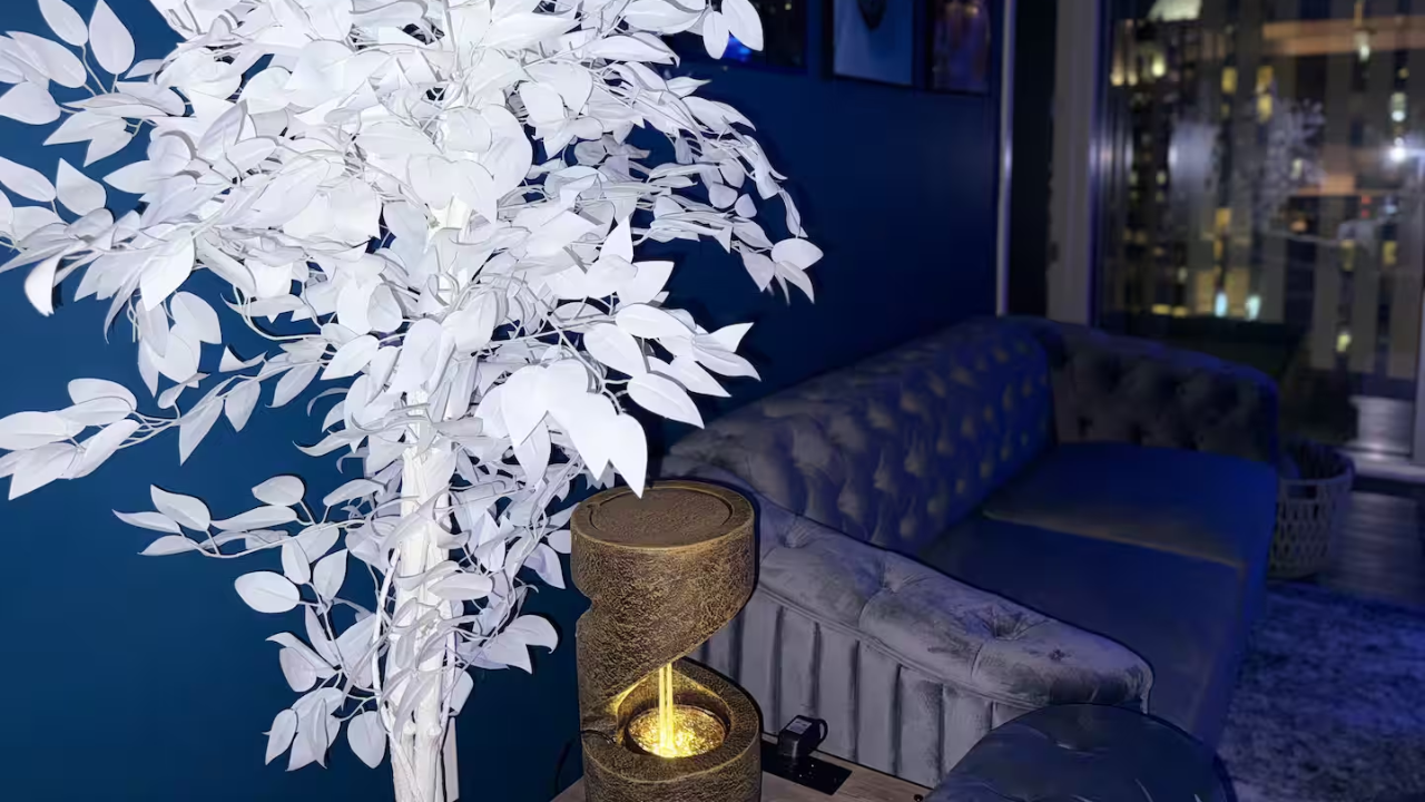 Interior of a modern living room with a white artificial tree, a vintage-style table lamp, a velvet sofa, and city lights visible through large windows.