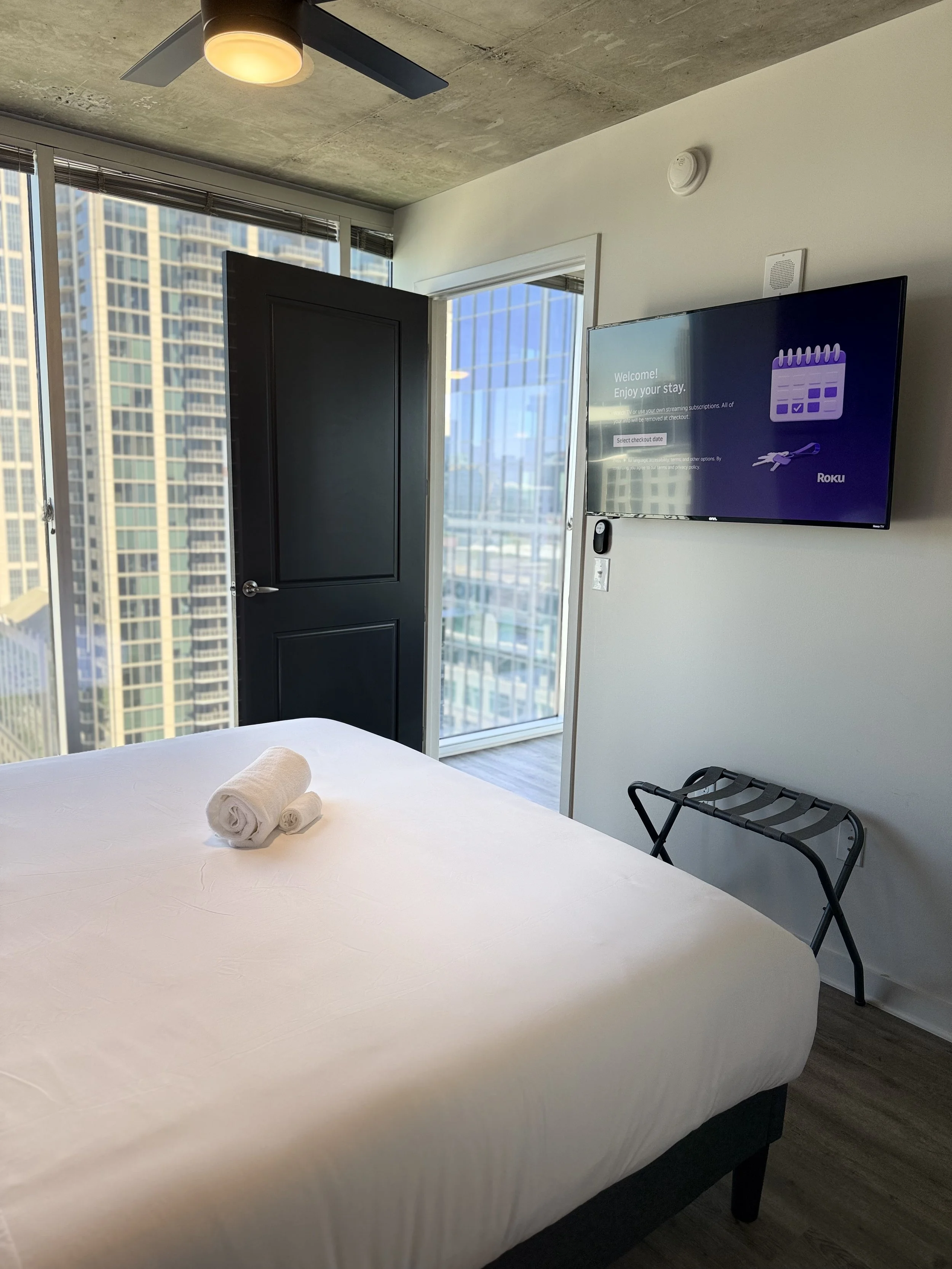 Modern hotel room with a neatly made bed, white towels on top, a black luggage rack, a wall-mounted television, a door open to a balcony with city views, and a ceiling fan with lighting.