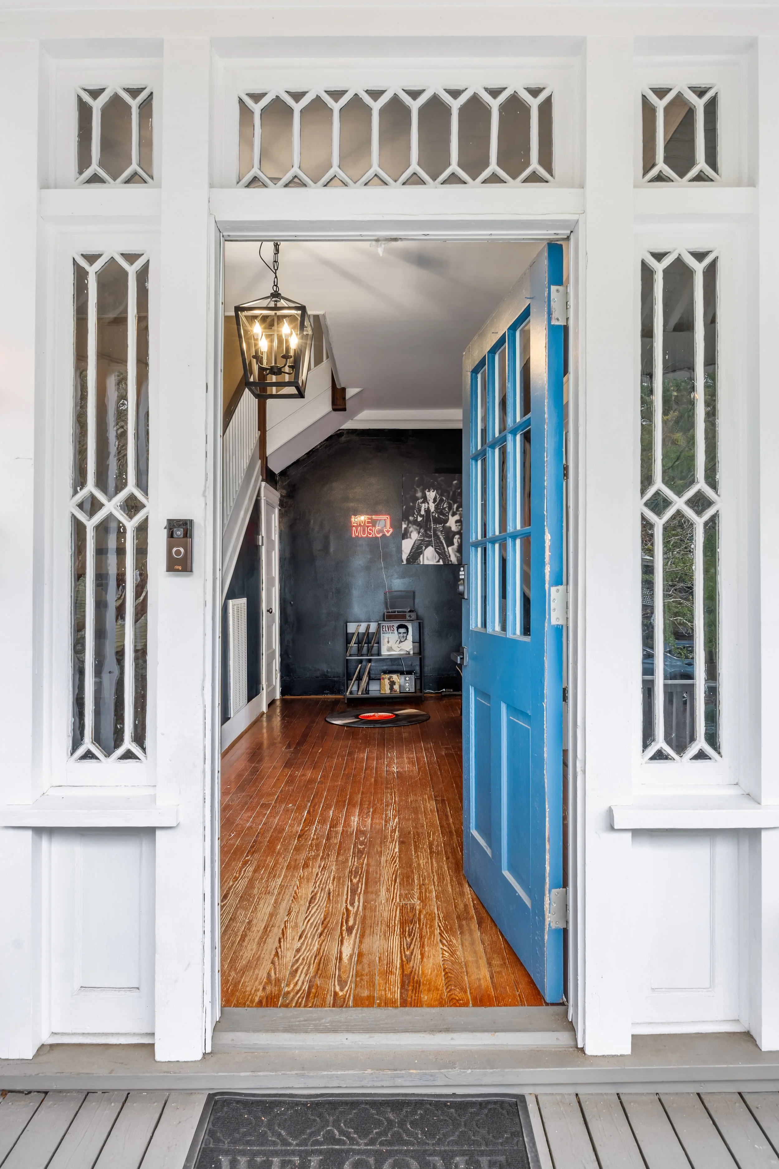 View through a front door into a room decorated with a music theme, featuring a black chalkboard wall with a neon sign that says 'Live Music', a small record shelf, and a vinyl record on the floor, with a blue door open to the porch.