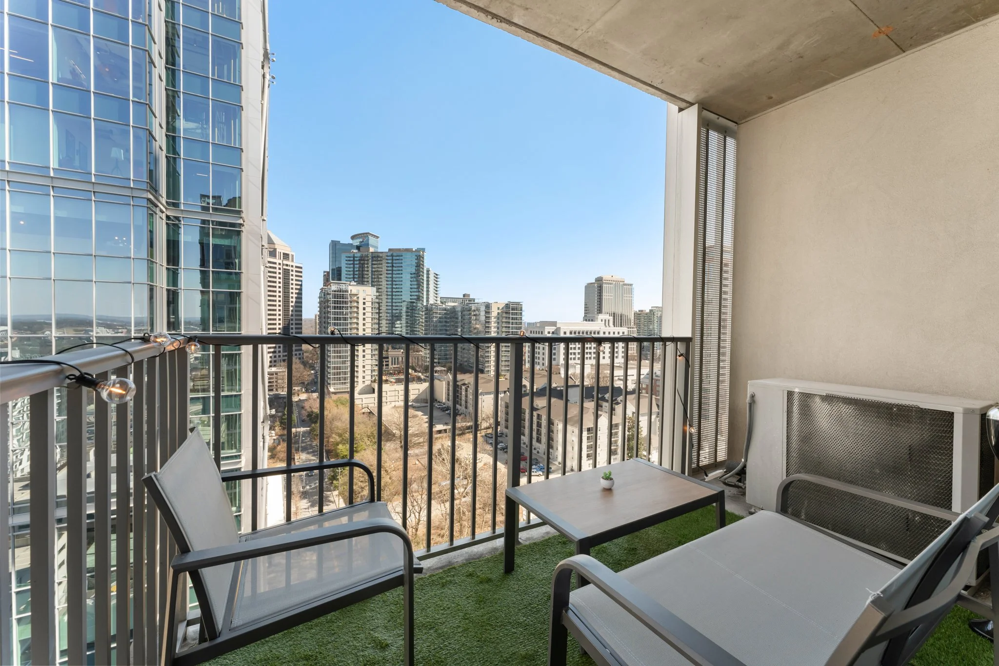 Small balcony with outdoor furniture, including two chairs and a table, overlooking a cityscape with tall buildings, under a clear blue sky.