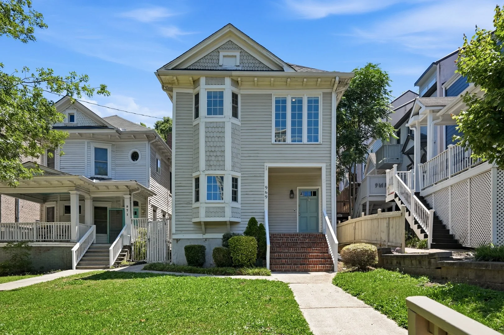 A two-story white house with a small front yard, bushes, and a brick staircase leading to a light blue front door. The house features bay windows and is surrounded by neighboring houses with stairs and decks, under a clear blue sky.
