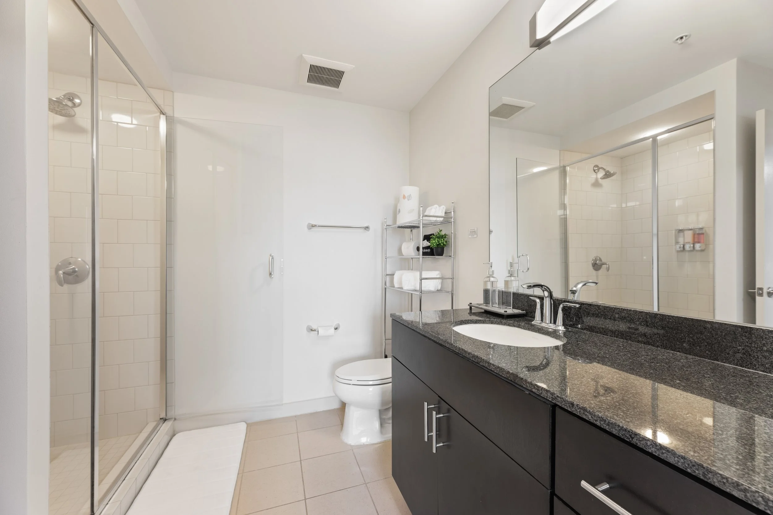 Modern bathroom with a glass-enclosed shower, white tiles, a white toilet, a gray countertop vanity with a large mirror, and shelves with towels and toiletries.
